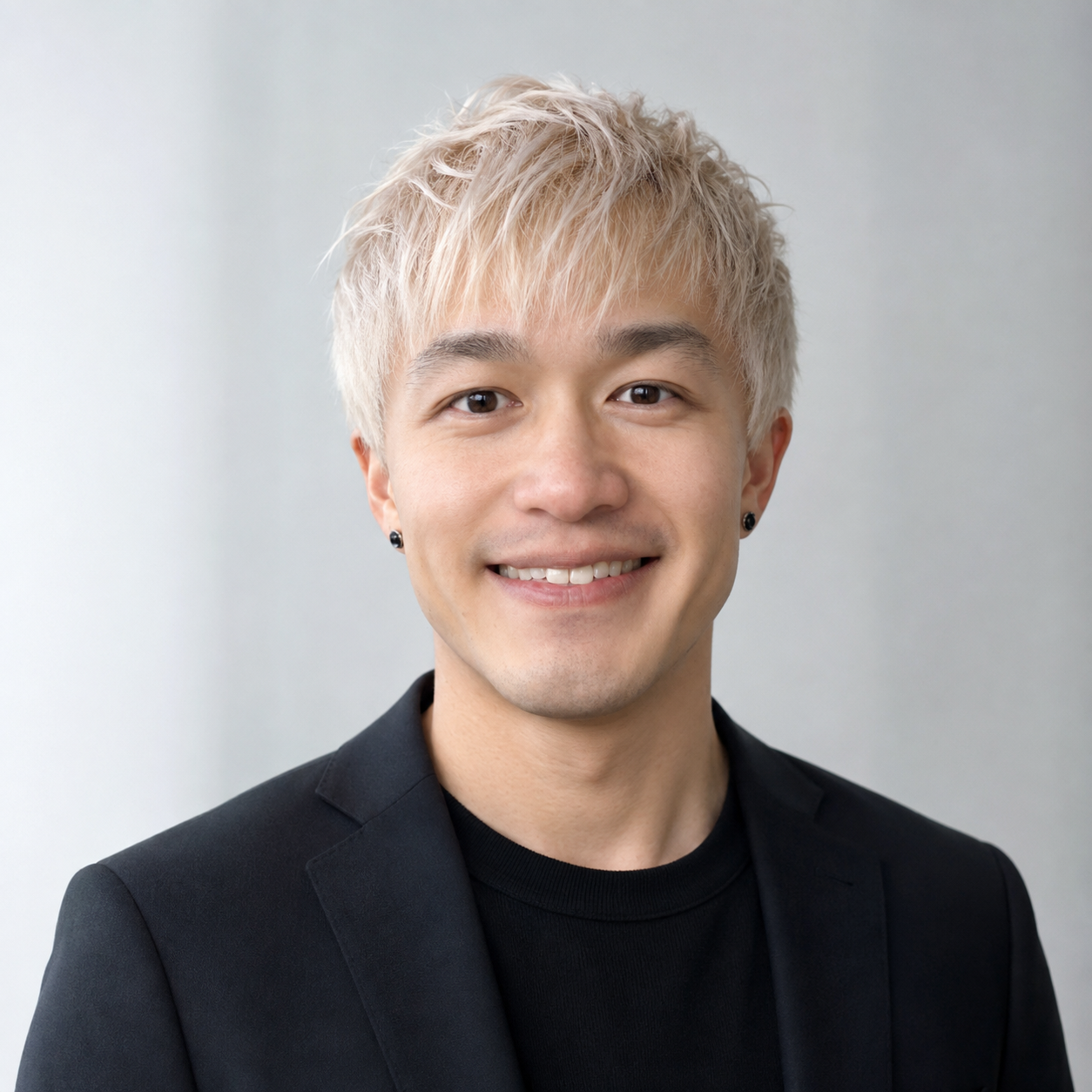 A young man with short, bleached blonde hair, wearing a black suit and black shirt, smiling at the camera against a plain light gray background.