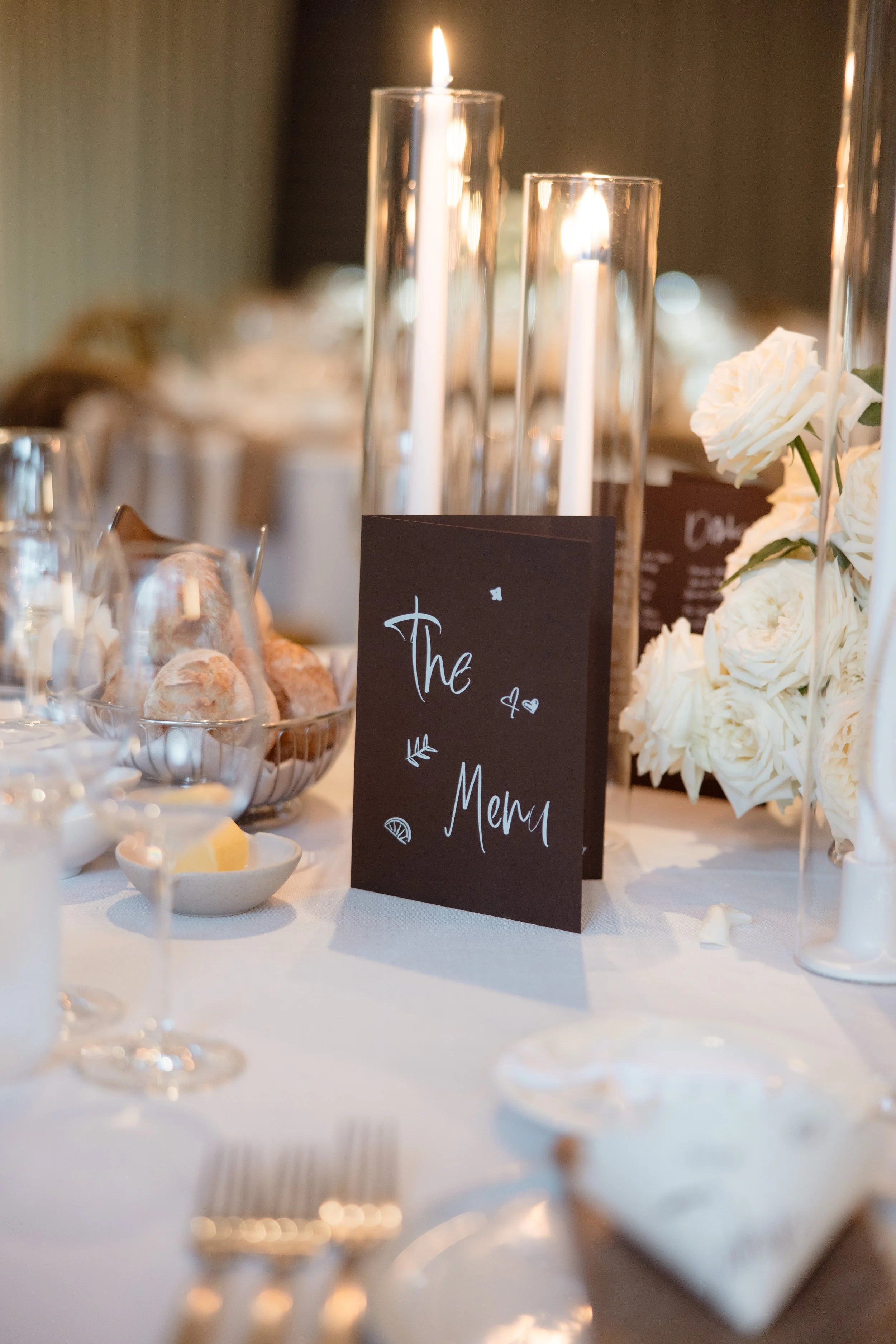 Wedding guest table styled by Philoxi Styling Melbourne. Features a mocha brown custom table menu, tall dinner candles in sleeves, white rose centrepiece , wine glasses, bread rolls, cutlery and crockery. Set and ready for the wedding reception. 