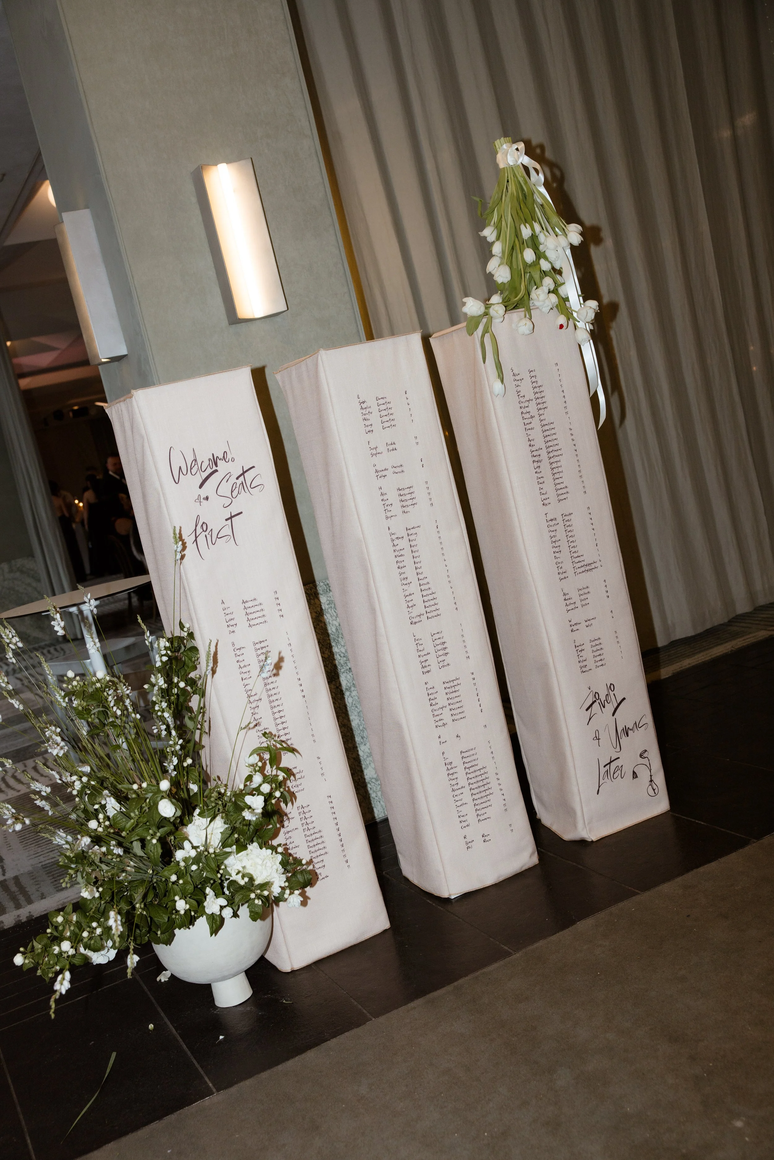 Wedding guest seating chart in the welcome foyer. Styled by Philoxi Styling Melbourne - three tall columns feature linen covers with guest names and table numbers printed. White tulips and a floor arrangement are used as decor. 