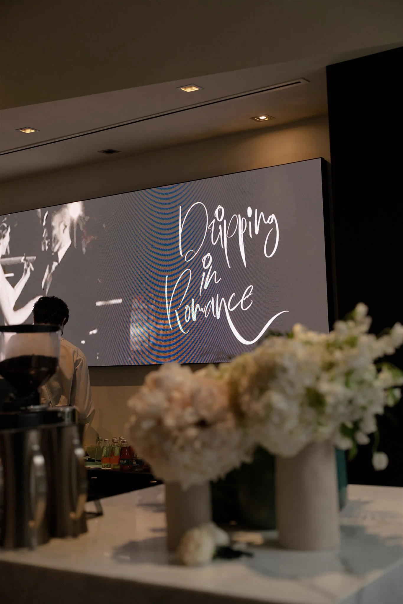 Image of a romantic wedding table setup. Pastel florals (tulips, roses and and greenery) in ceramic vases, blush dinner candles, brushed gold lamp and textured napkins adorn the table. 