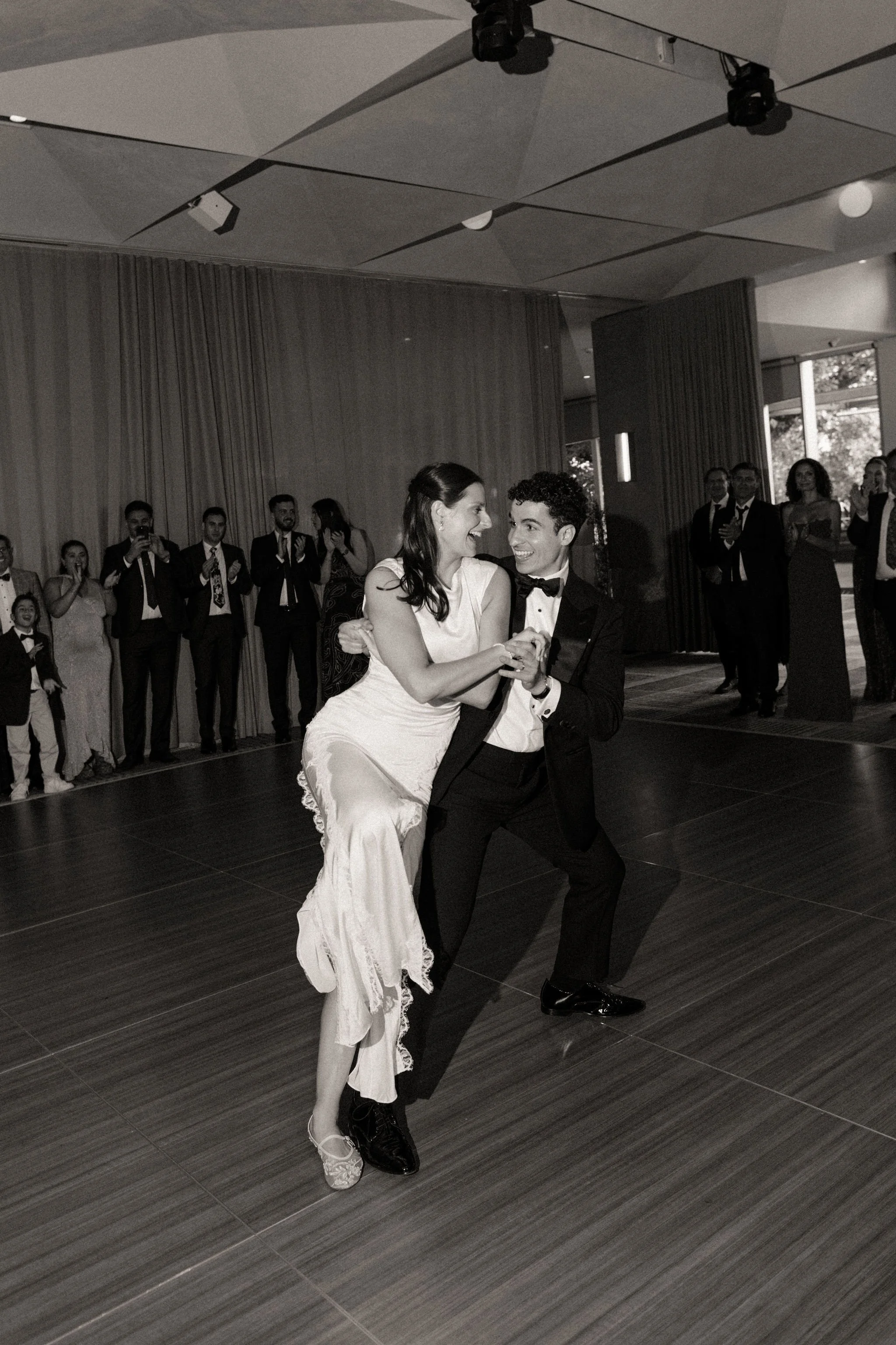 Couple's first dance. The Bride leans into the Groom as he dips her to he side. Guests around the dance floor clap as they watch the first dance. Bride wears a sleek, modern Shona Joy Wedding Dress as her second dress.