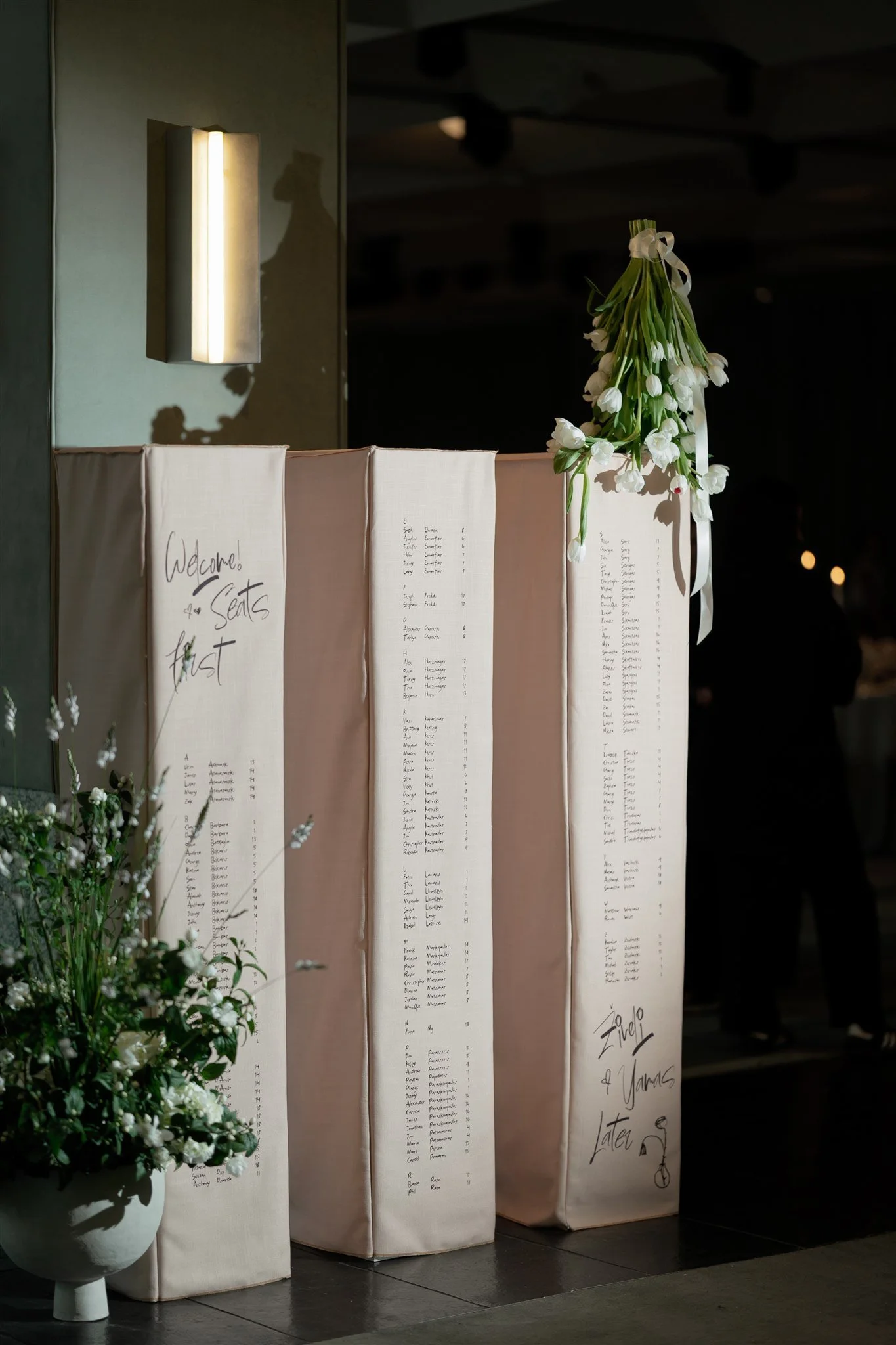 Wedding guest seating chart in the welcome foyer. Styled by Philoxi Styling Melbourne - three tall columns feature linen covers with guest names and table numbers printed. White tulips and a floor arrangement are used as decor. 