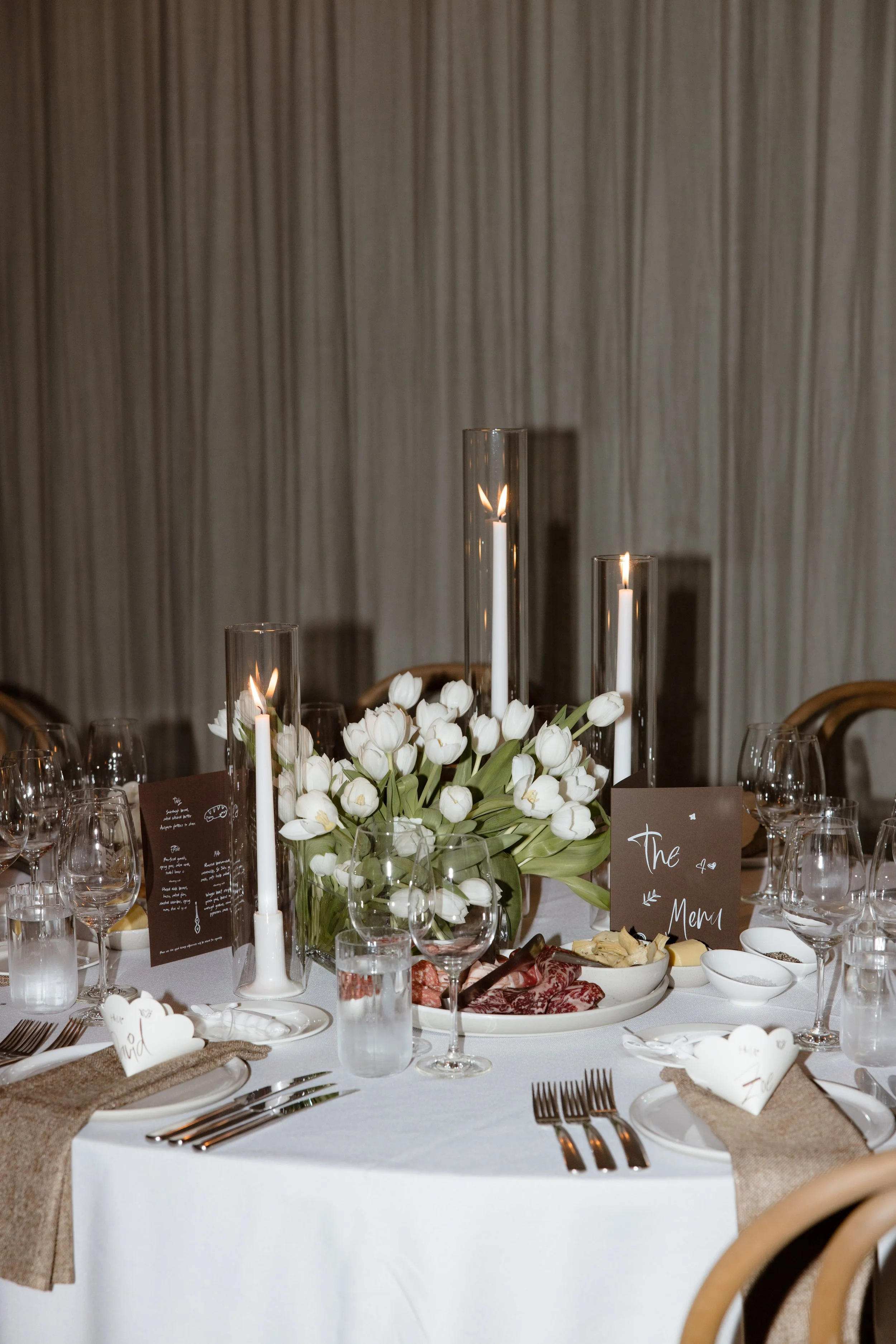 Image by Theodore and Co, Melbourne Wedding Photographers - featuring a beautifully styled guest table with cream linen, brown textured napkins, Bentwood chairs, vintage-inspired placecards and table menus, white tulips, candlelight. Antipasto Plate.