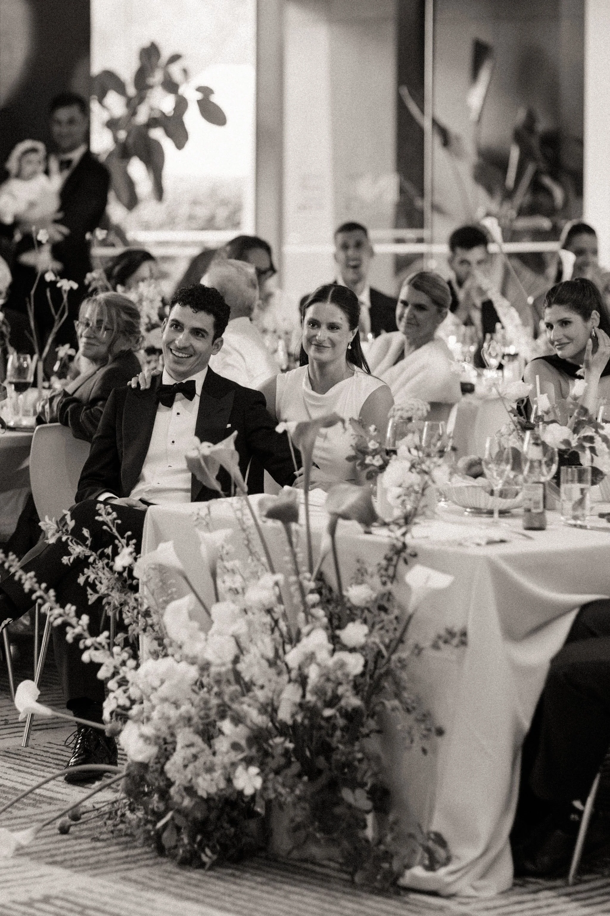 Black and White photo of Bride and Groom sitting at long dining table amongst their guests. A large arrangement of classic green and white florals sits at on the floor at the end of the table. 
