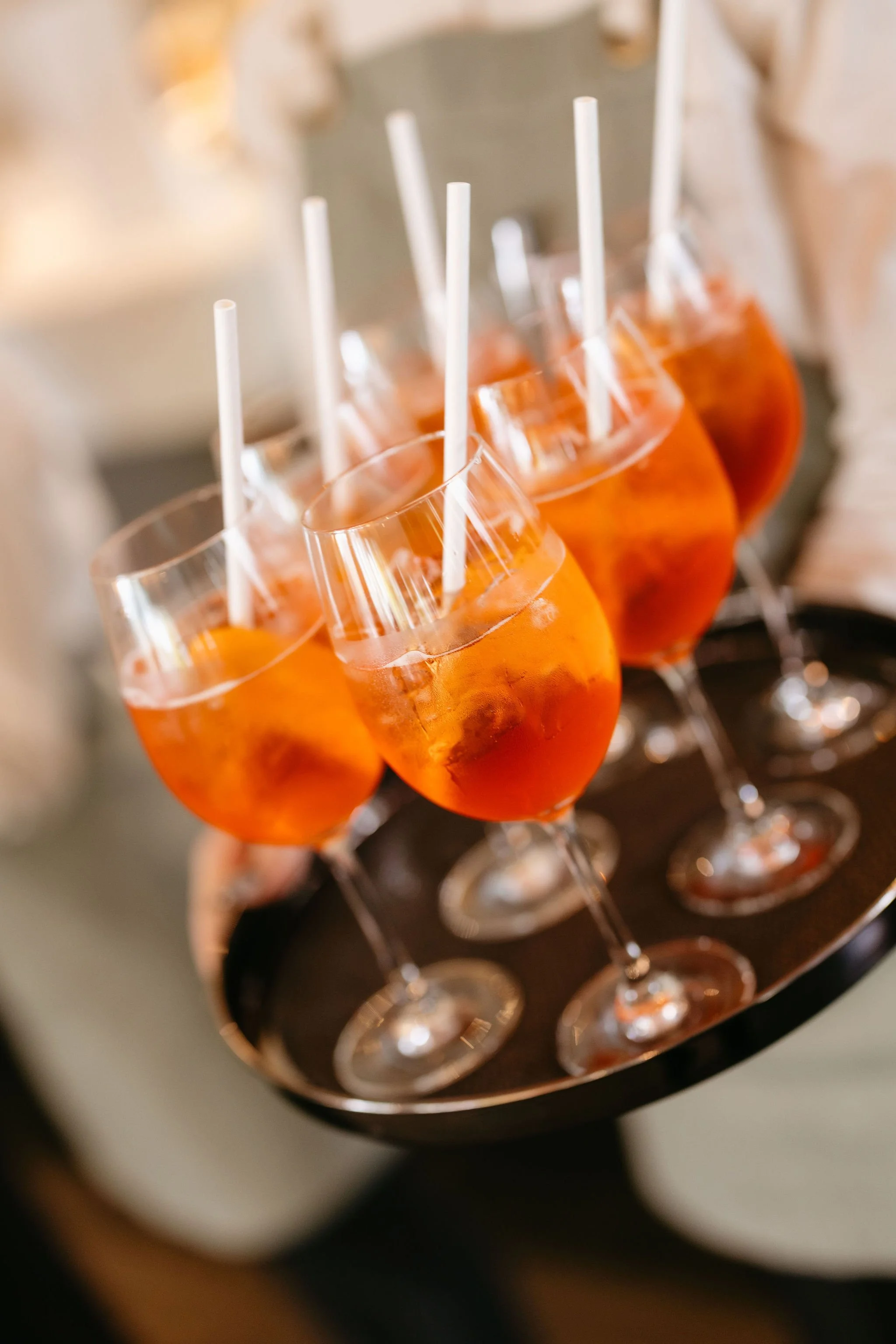 Image shows a waiter holding a round chrome tray with six Negroni cocktail in wine glasses being served to wedding guests during cocktail hour/ pre-dinner drinks. 