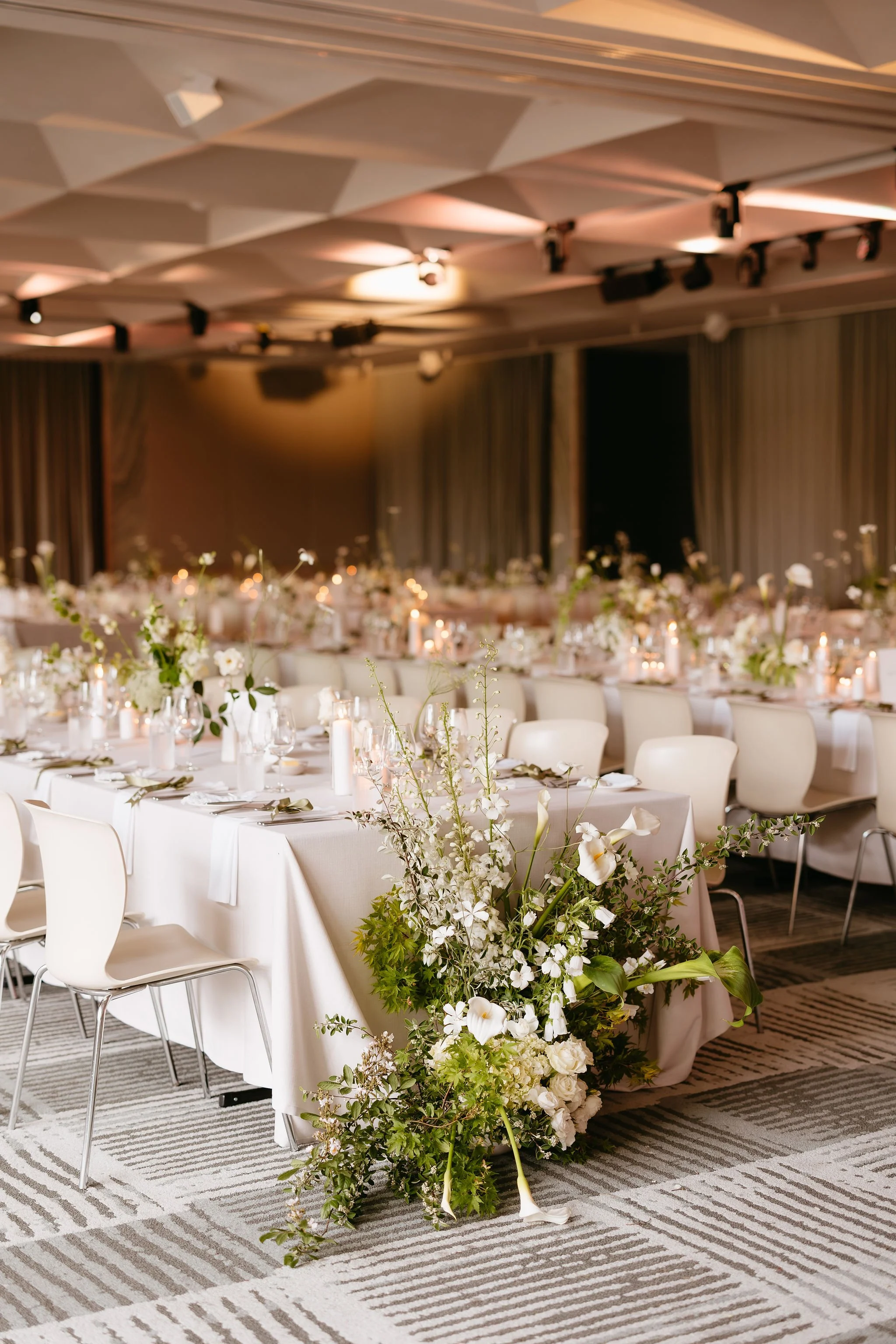 Image of long dining table setting at a wedding. Cream textured linen, single stem flowers in bud vases, and candlelight adorn the tables giving a sleek, minimalistic vibe. The end of each table features a large floor arrangement of sculptural floral