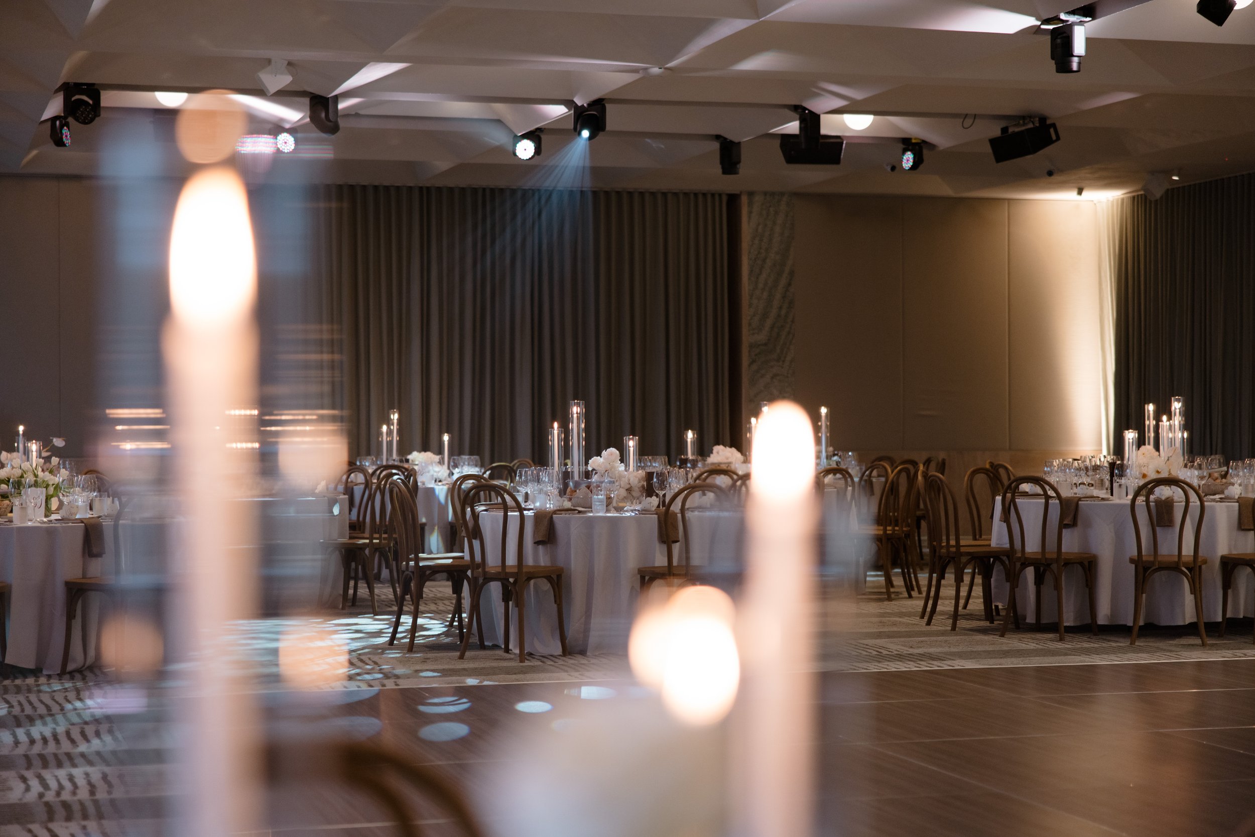 Contemporary wedding setup at Zinc at Fed Square. Round tables styled with warm neutrals - offwhite textured tableclothes, brown napkins, vintage-inspired stationery details and stunning, low, organic floral centrepieces surrounded by tall candles