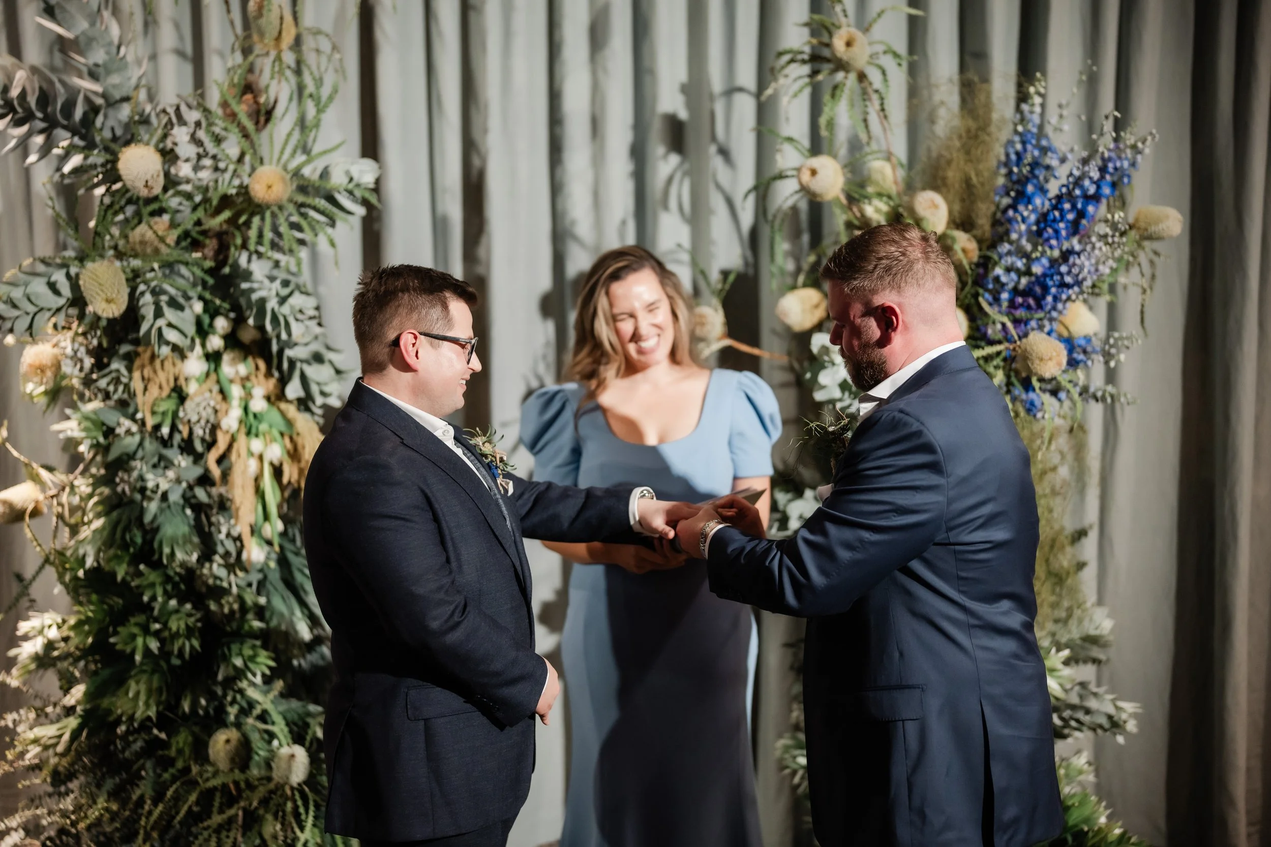Same-sex couple civil ceremony taking place in Studio 1 and Zinc. Groom and Groom in navy wedding suits exchanging wedding rings and vows. Celebrant is wearing a light blue cocktail dress. Structural florals sit in front of a softly draped wall.  