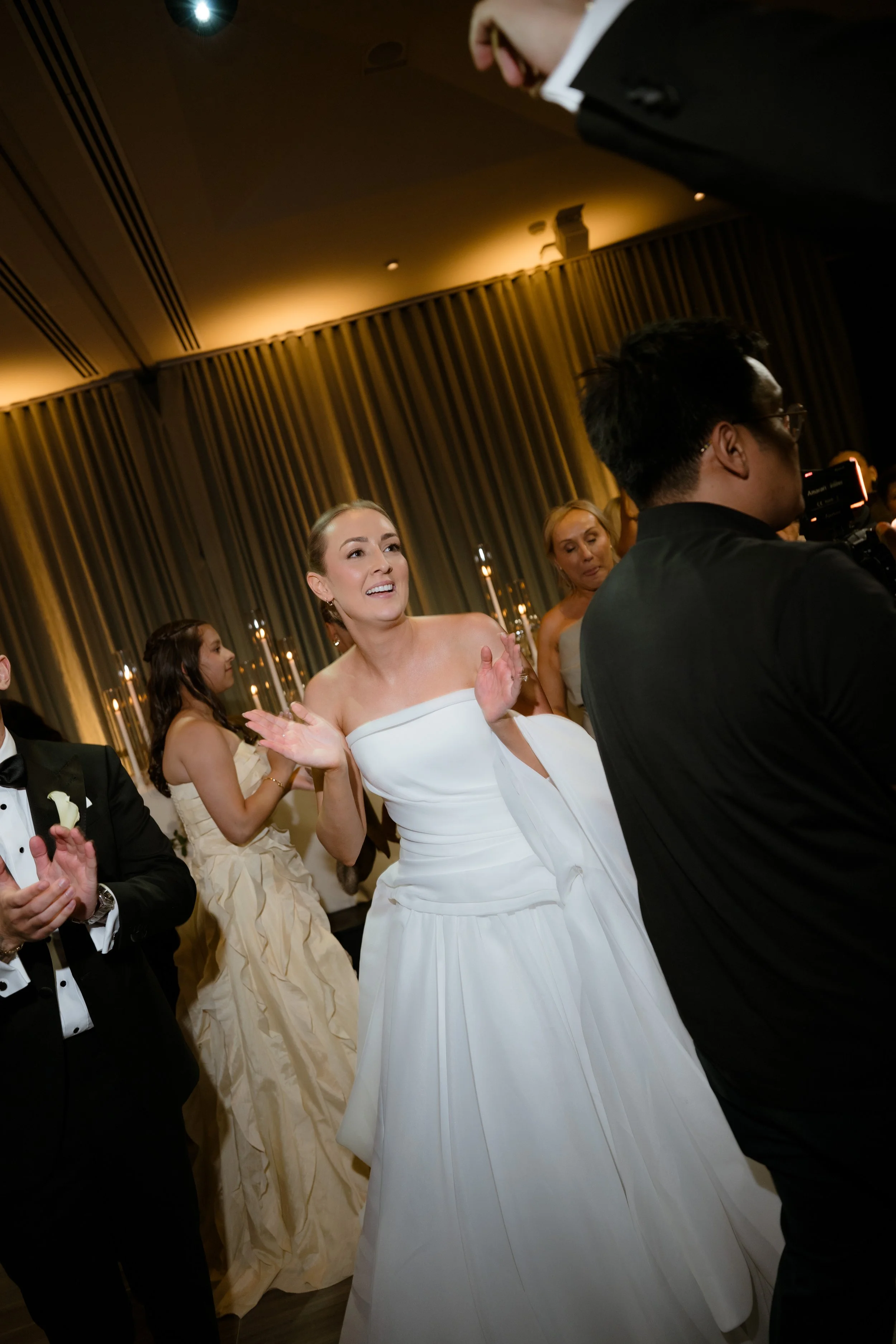 Bride dancing at wedding reception. She wears a contemporary strapless off-white wedding gown. Guests surround her wearing black tie attire. Candlelight glows in the background. 