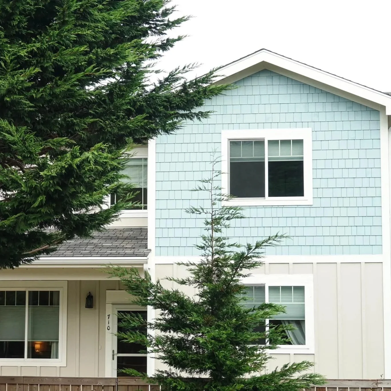 A two-story house with light blue siding, large windows, and a large evergreen tree in front partially obscuring the view.