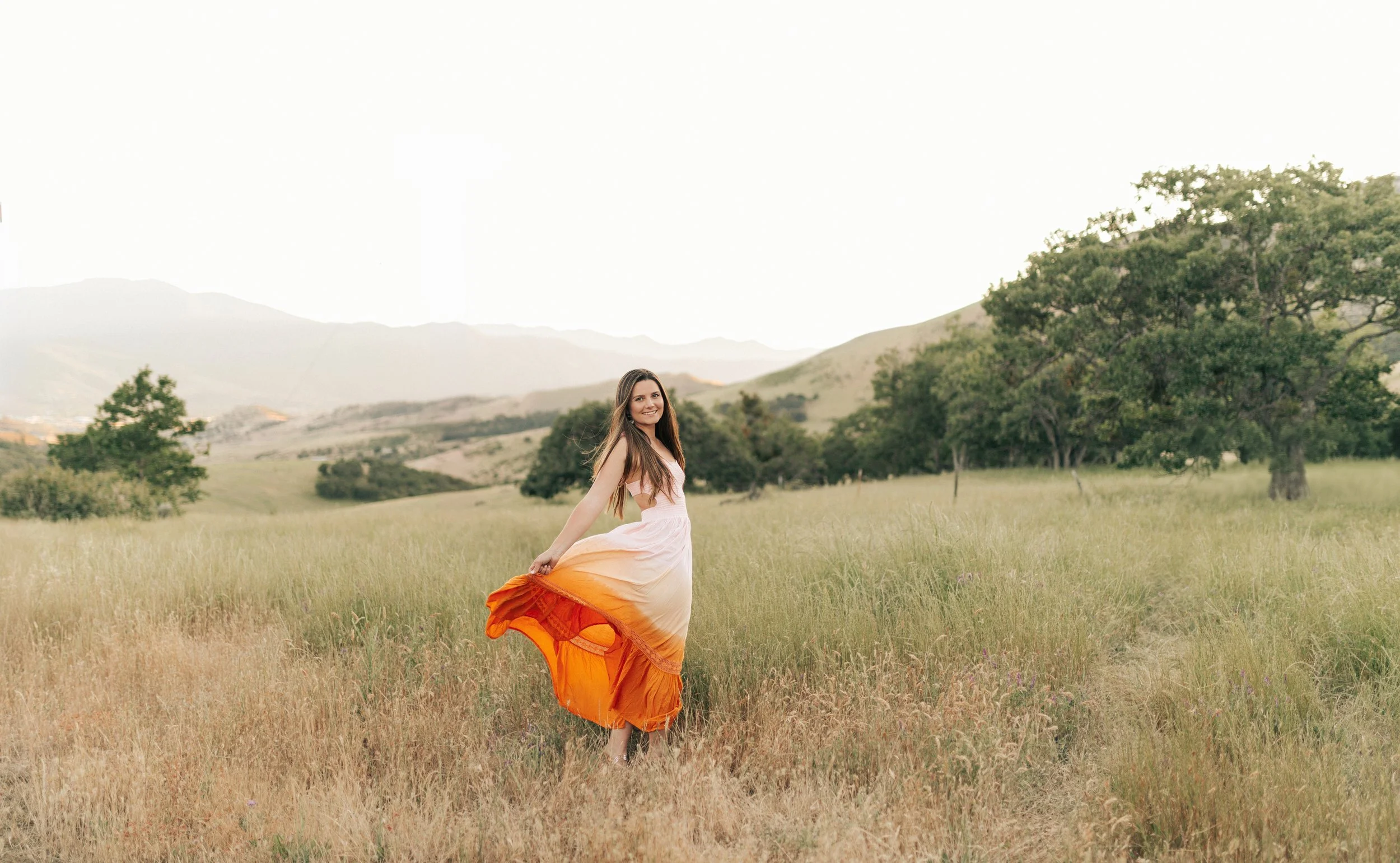 A young woman, Alexis Willick, in a flowing pink and orange dress standing in a grassy field with trees and mountains in the background in southern oregon.