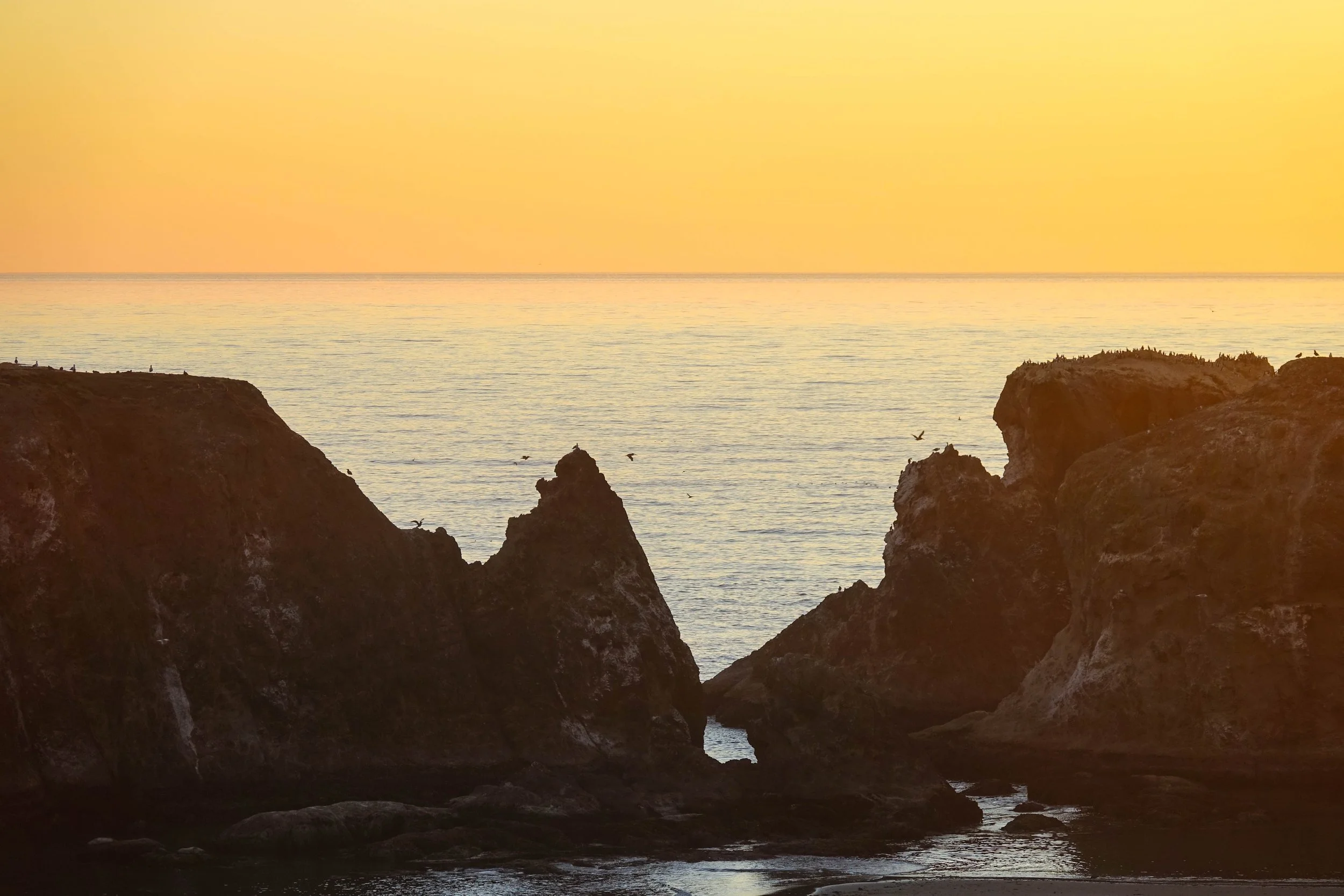 A coastal scene at sunset with cliffs, calm ocean, and a yellow-orange sky , taken by Bandon, Oregon Realtor.