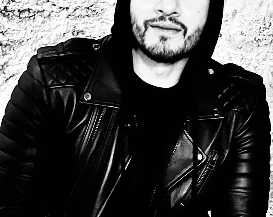 A man with a beard wearing a hooded jacket and leather jacket sitting against a textured wall.