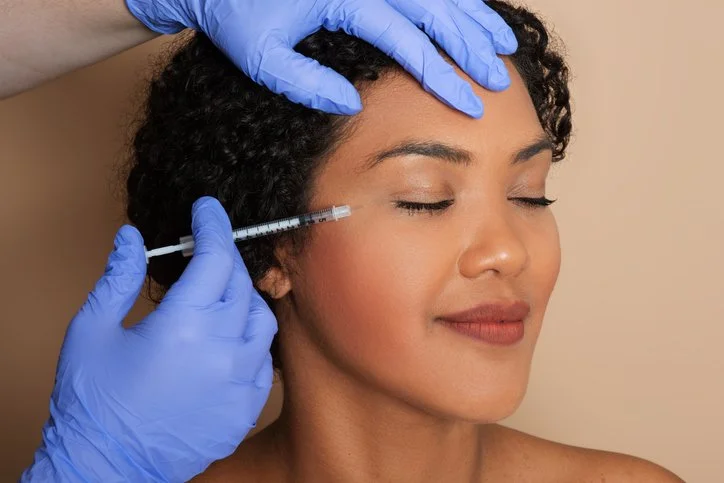 A woman with curly hair receives a cosmetic injection in her forehead from a medical professional wearing blue gloves.
