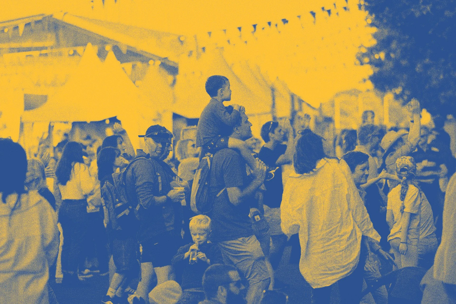 Crowd of people at an outdoor event or festival during the evening, with tents and string lights in the background.