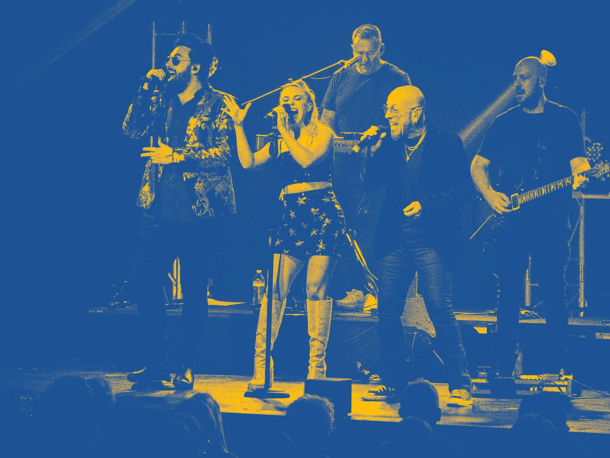 A group of five musicians performing on stage, with two women and three men, one holding a microphone, another playing an electric guitar, and others singing or playing instruments, with an audience in front.