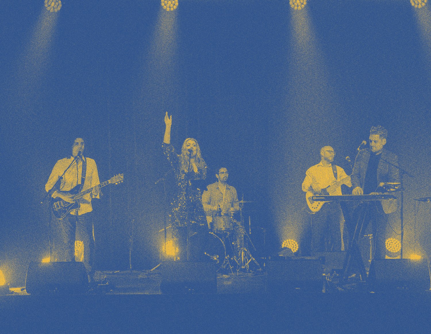 A band of five musicians performing on a stage, including a female singer, two guitarists, a drummer, and a keyboardist, illuminated by stage lights.