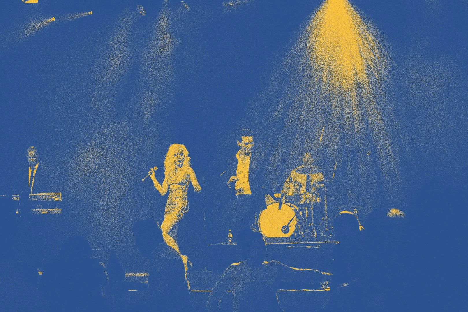 A live music performance with a female singer, a male singer, and a drummer on stage, illuminated by yellow stage lights.