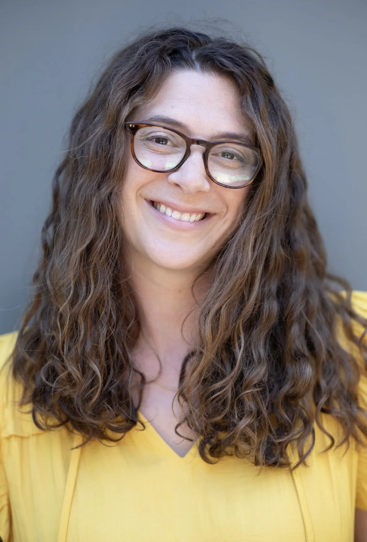 A woman with long, curly brown hair and glasses smiling at the camera, wearing a yellow top, standing against a gray background.