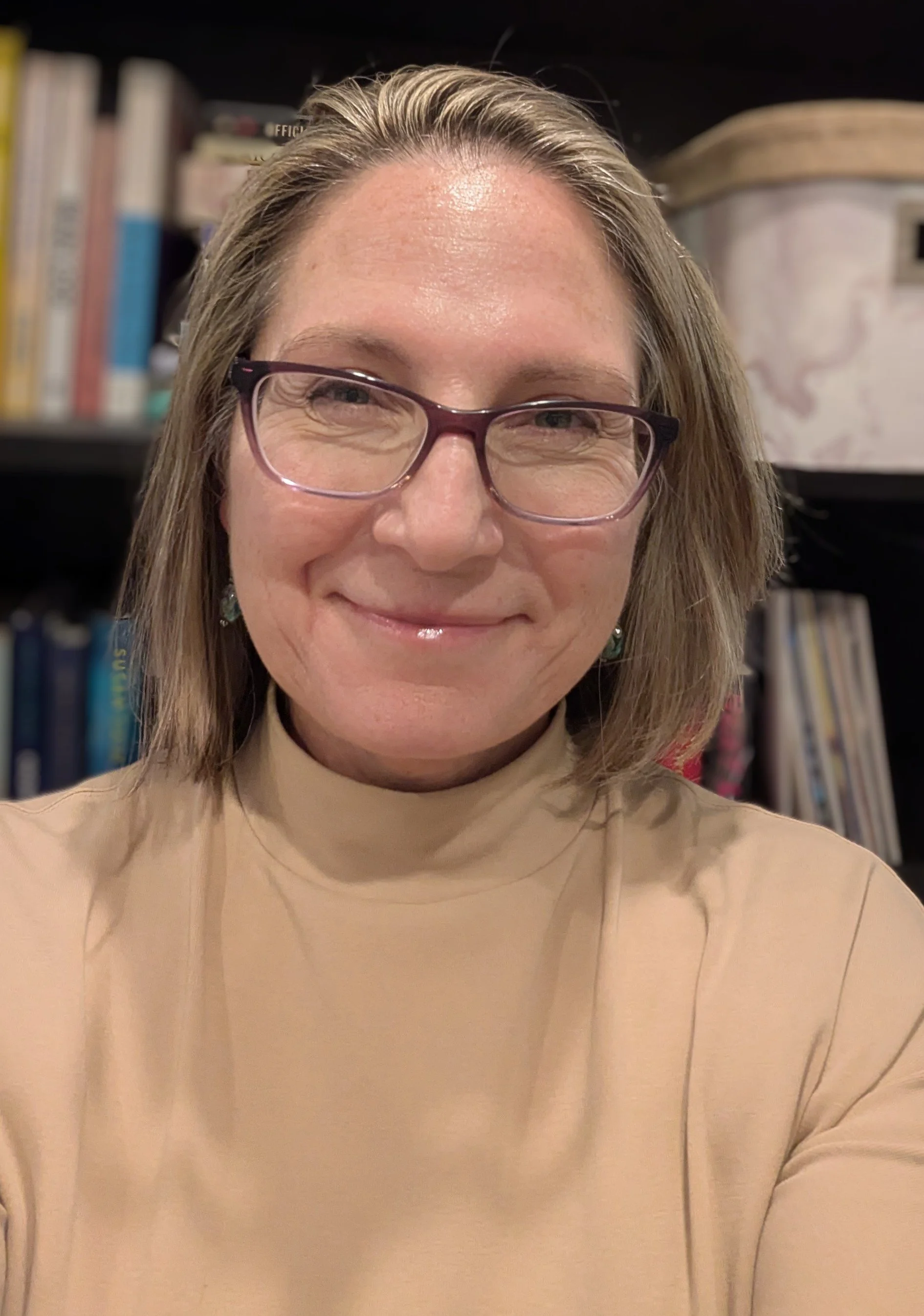 A woman smiling, wearing glasses and a beige top, with bookshelves in the background.