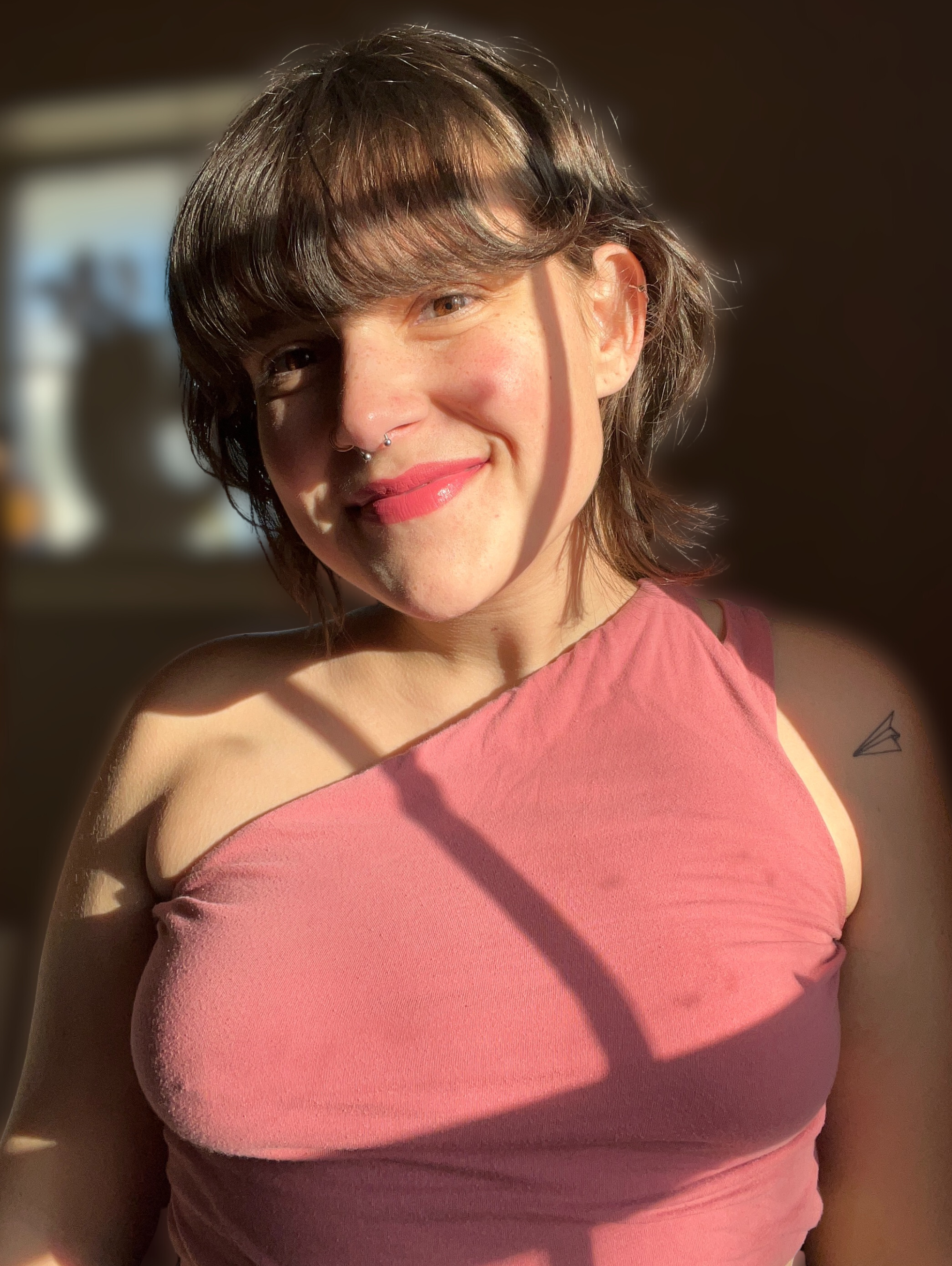 A young woman with short, wavy brown hair, wearing a pink top, smiling in natural sunlight, with a shadow across her face.