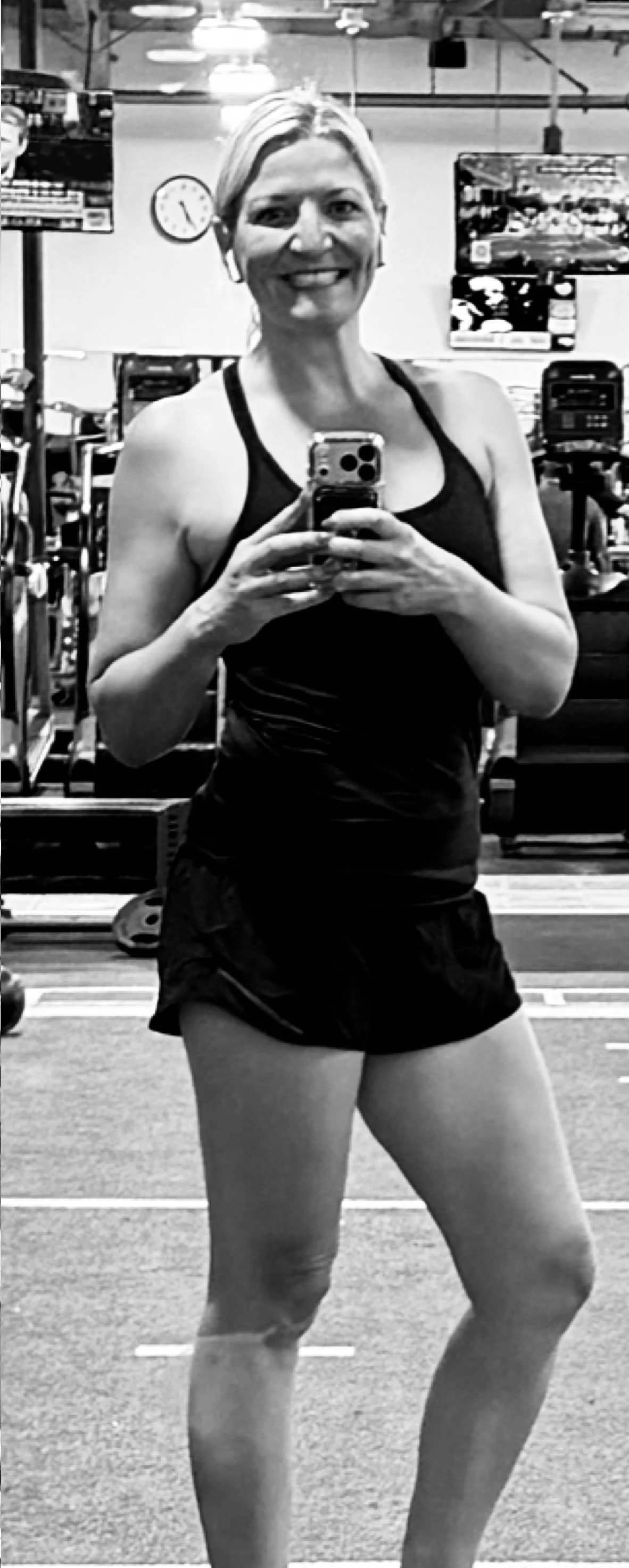 Woman taking a selfie in a gym mirror, smiling, wearing a black tank top and shorts.