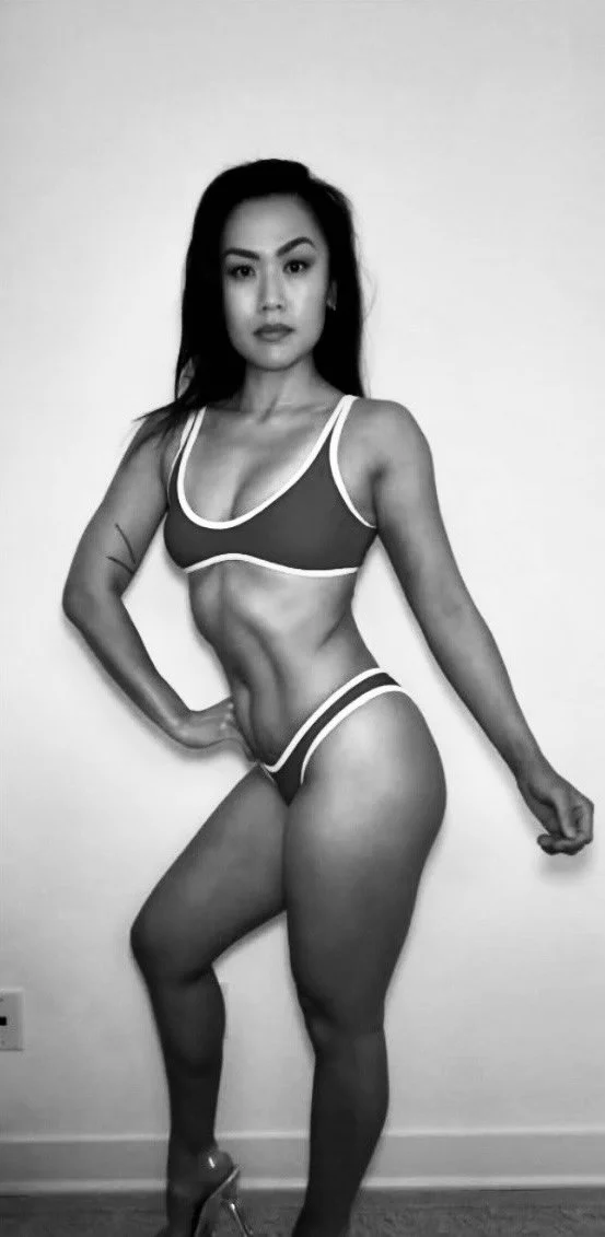 A woman with long dark hair in a sports bra and shorts, striking a pose against a plain wall.