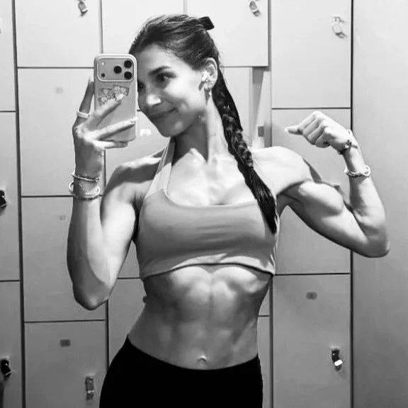A woman in workout clothes lifting weights in a gym, smiling, with a person stretching on the floor in the background.