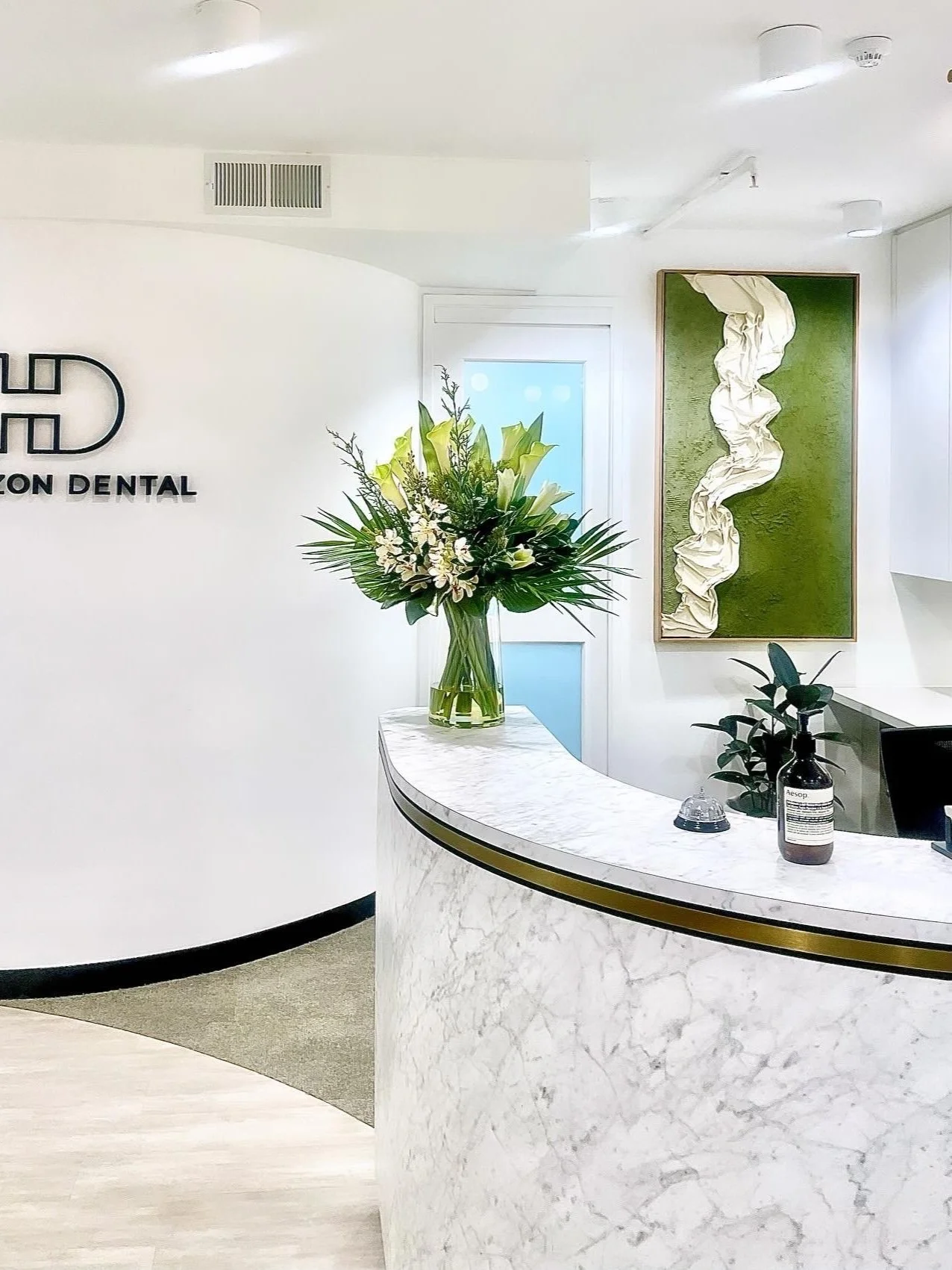 Reception area of a dental office with a large floral arrangement on a white marble counter, a green plant, and wall art behind.