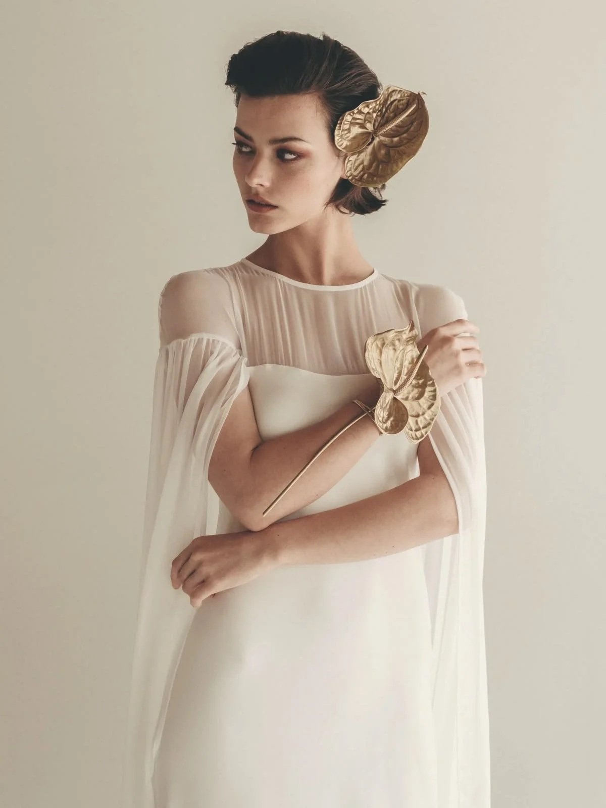 A woman with short dark hair wearing a white dress with sheer sleeves, holding and wearing gold leaf-shaped accessories.
