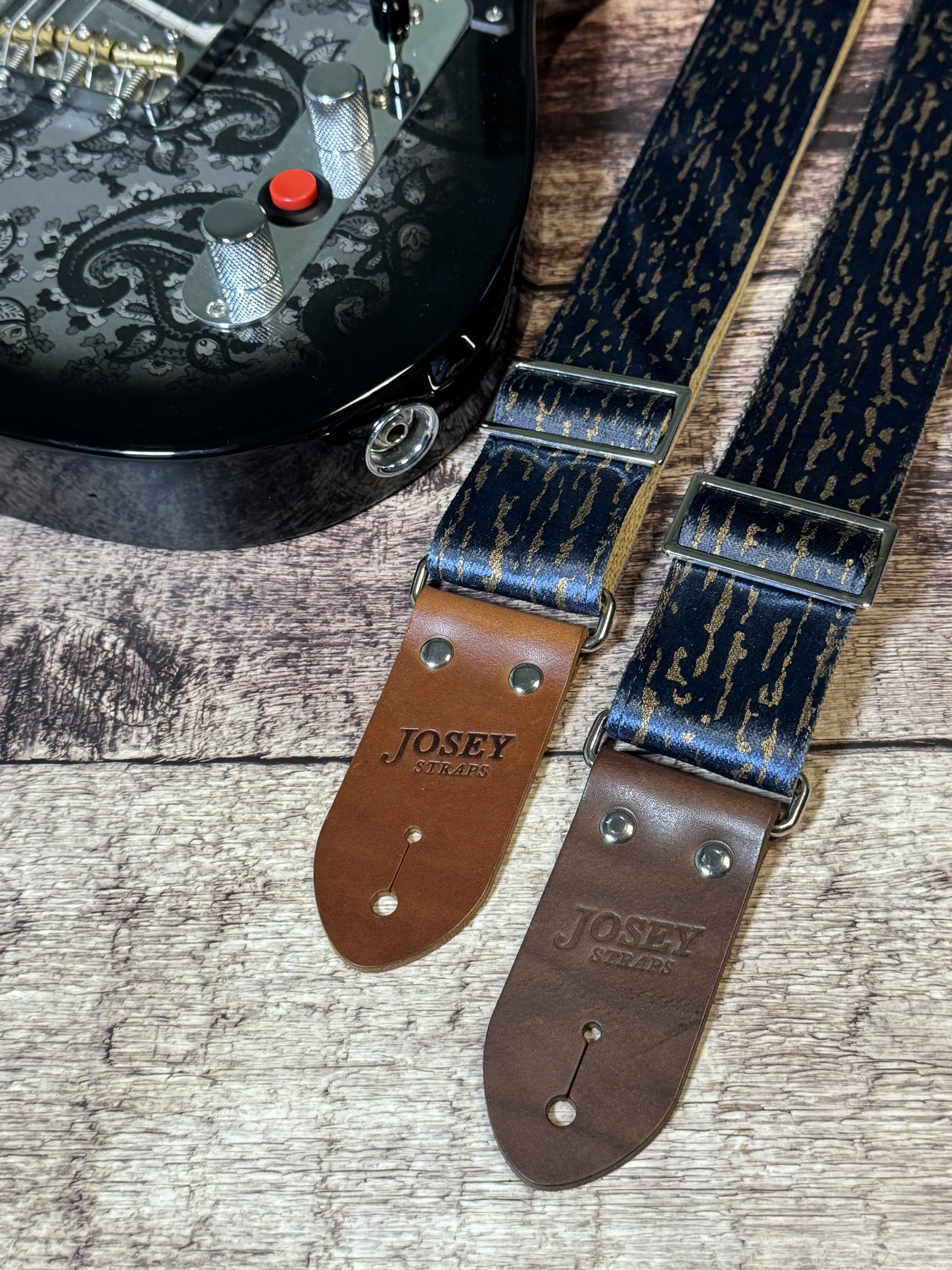 Blue and gold guitar straps on a wooden floor backdrop with a black paisley guitar in the background.