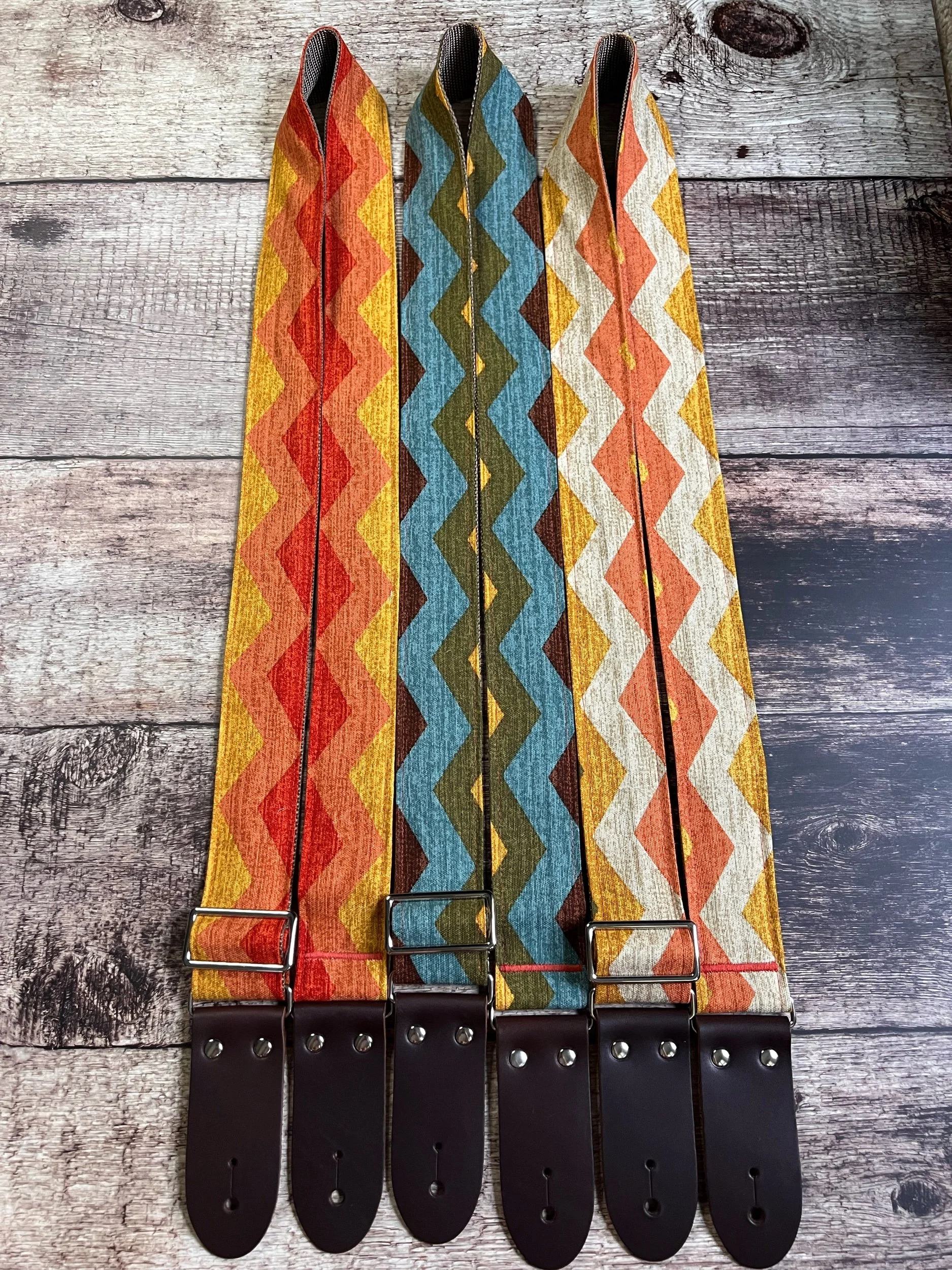 Four fabric guitar straps with colorful zigzag patterns in red, blue, white, and yellow, attached to black leather ends, laid out on a rustic wooden background.