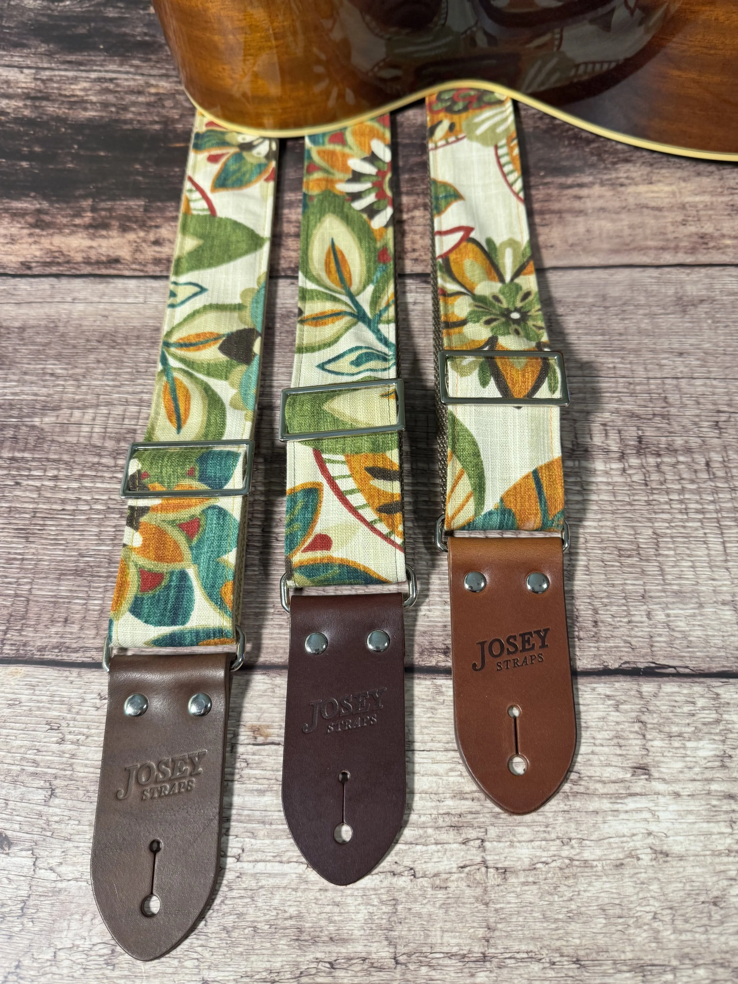 Handmade guitar strap in cream botanical fabric with amber, teal, burgundy, and forest green floral print, laid flat on a wood surface.