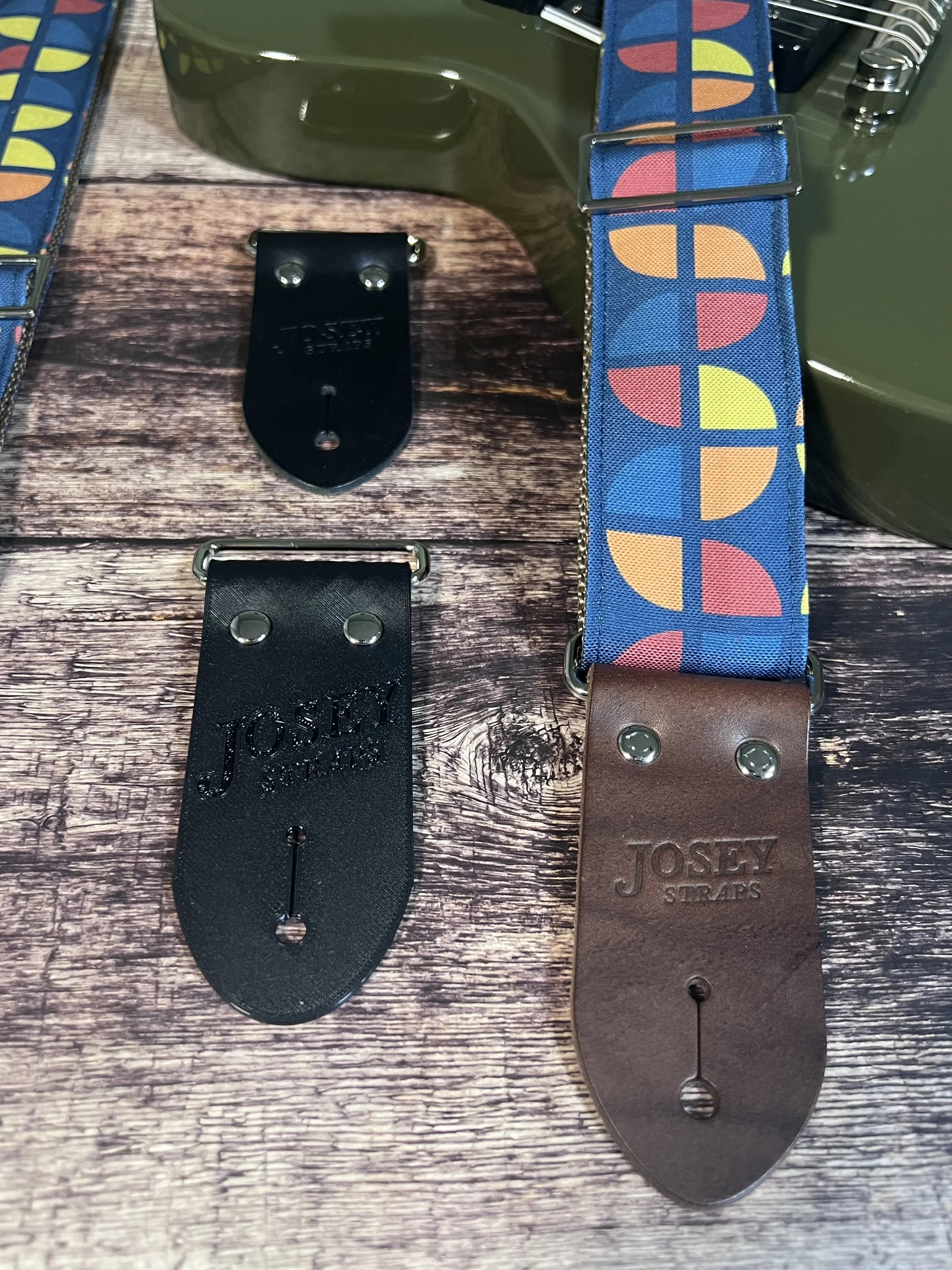 Two guitar end options in black, laying next to a navy guitar strap with geometric circles in mustard yellow, coral and steel blue with a brown leather Josey Straps end