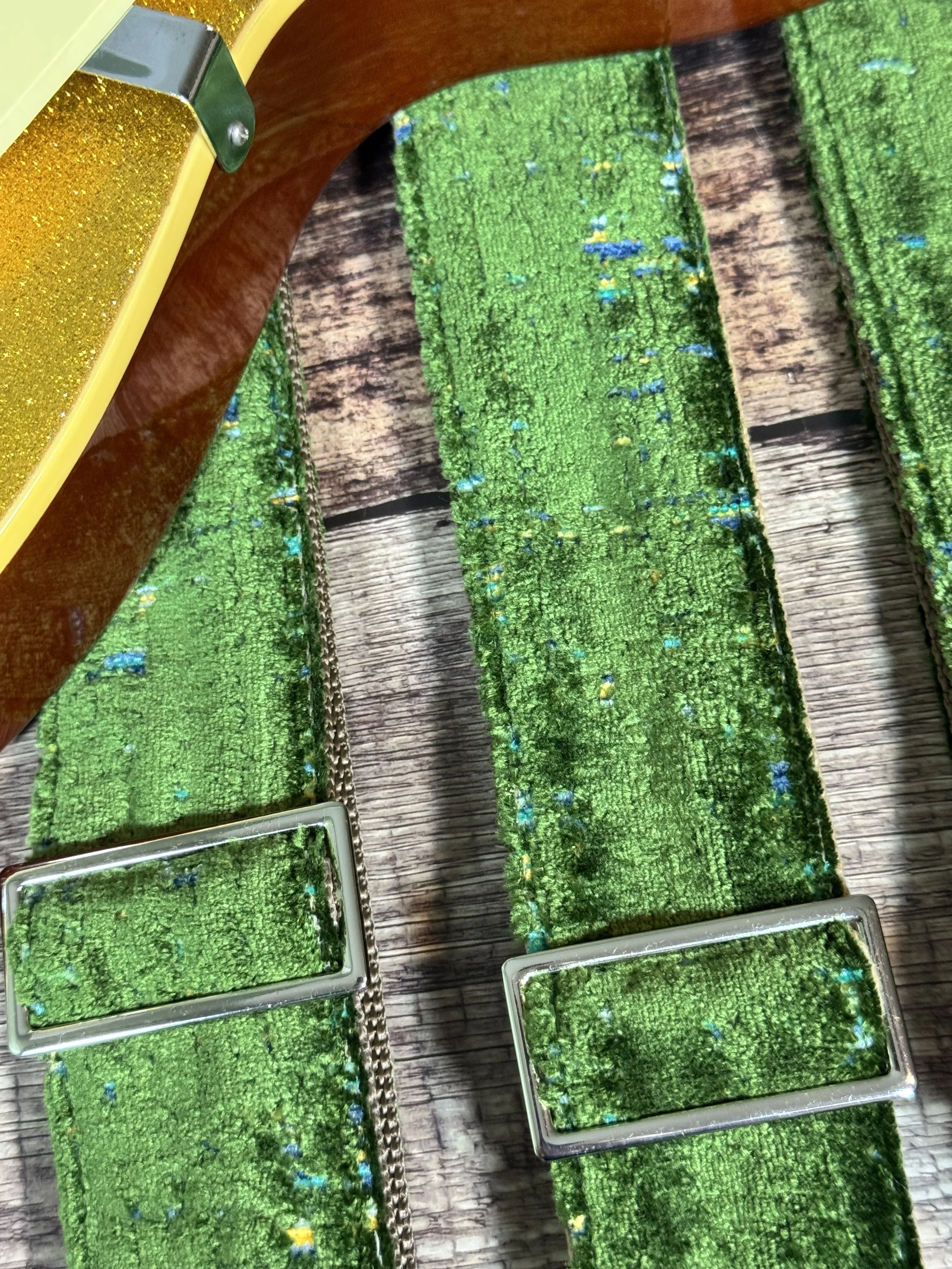Handmade green velvety guitar strap with stamped leather ends laid on weathered wood next to a gold sparkle electric guitar — The Grouch by Josey Straps