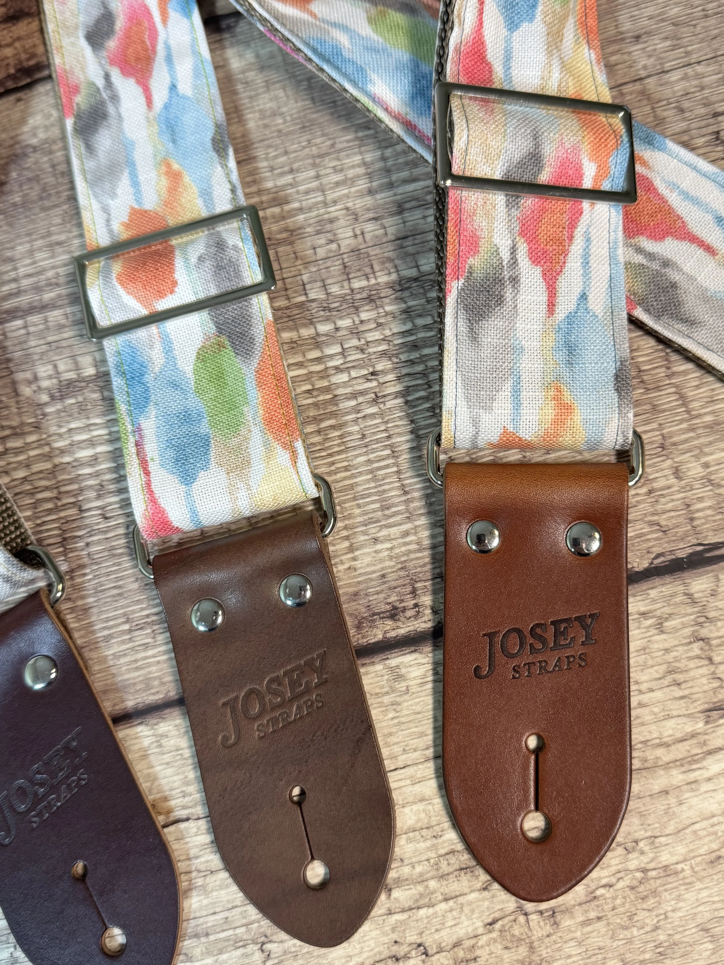 Josey Straps guitar straps in the April colorway — a watercolor print of coral, blue, sage, and amber on cream — displayed on a wood surface.