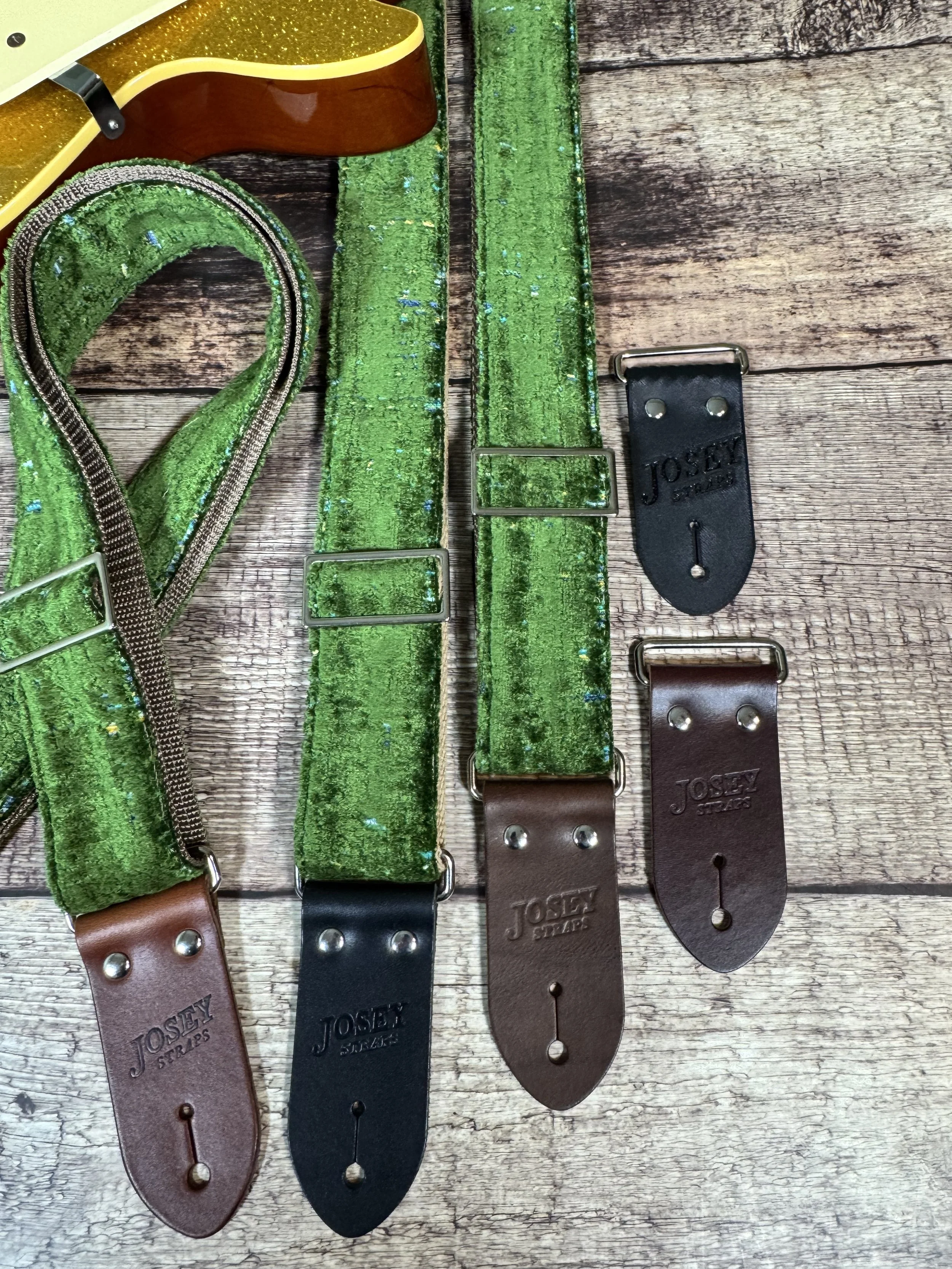 Handmade green velvety guitar strap with stamped leather ends laid on weathered wood next to a gold sparkle electric guitar — The Grouch by Josey Straps