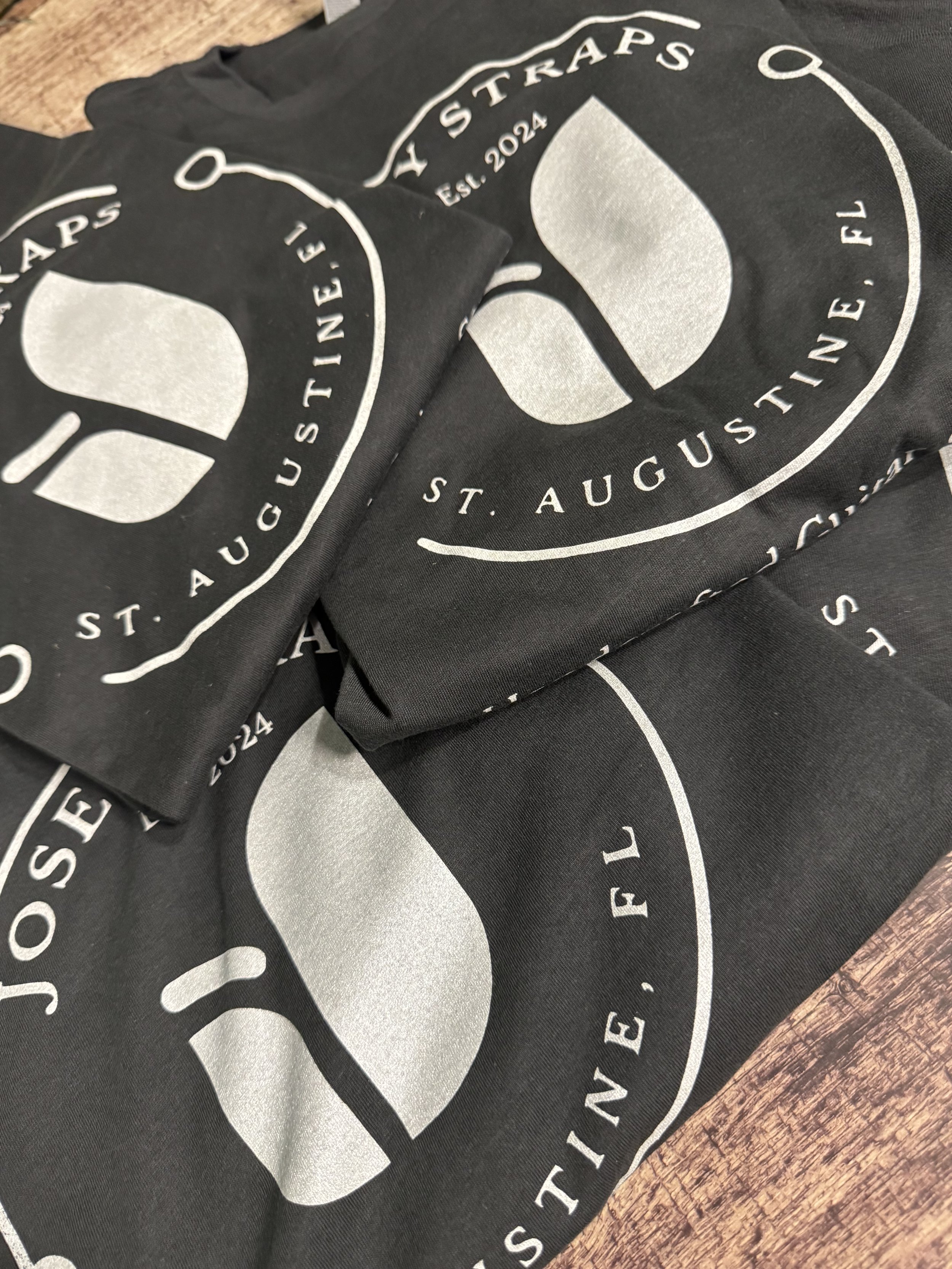 Multiple Josey Straps vintage black t-shirts arranged on a wooden floor backdrop.