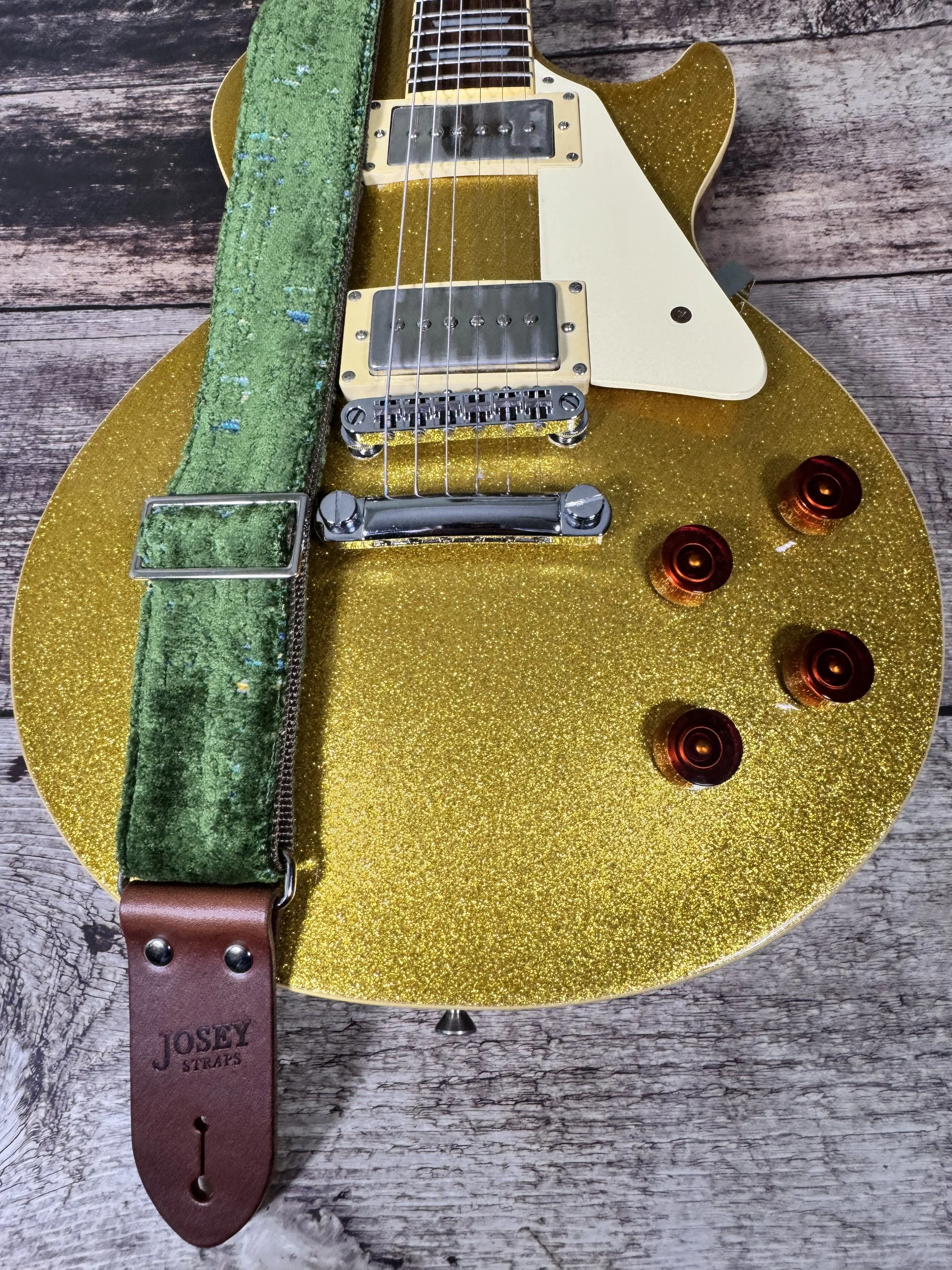 Handmade green velvety guitar strap with stamped leather ends laid on weathered wood next to a gold sparkle electric guitar — The Grouch by Josey Straps