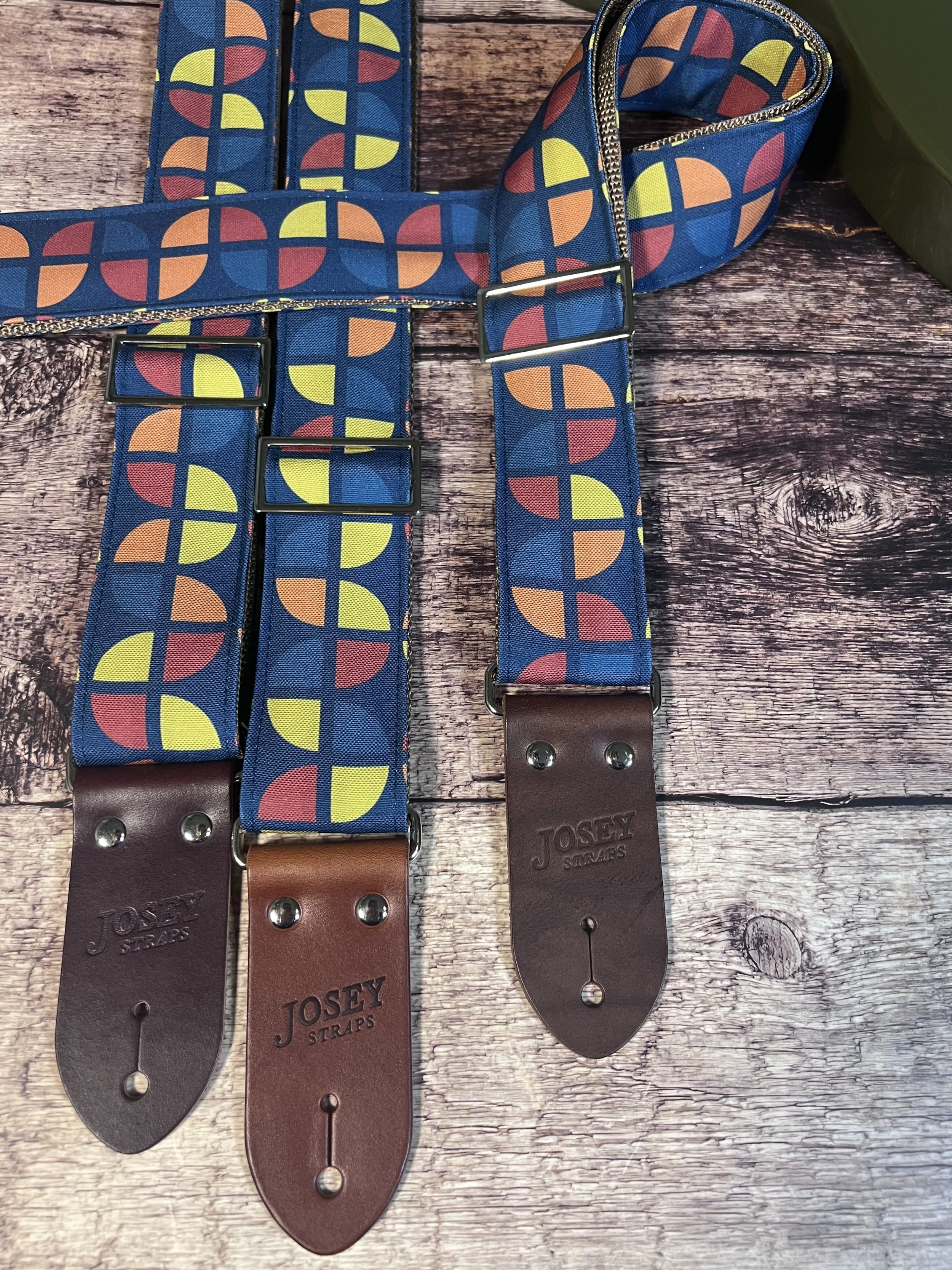 Three navy guitar straps, one folded, with groovy geometric circle pattern in mustard yellow, coral and steel blue
