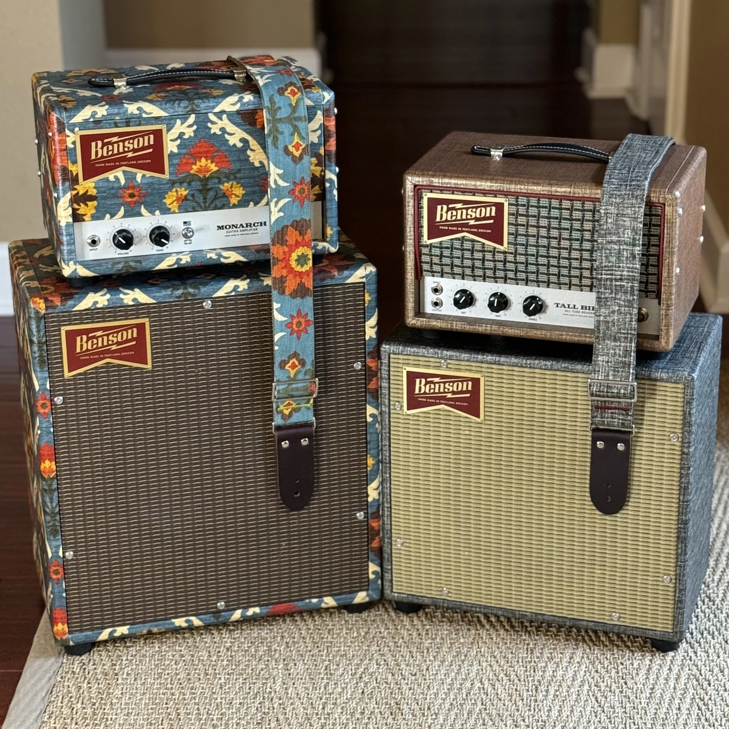 Four vintage Benson guitar amplifiers with patterned fabric coverings and straps, placed on a beige surface indoors.