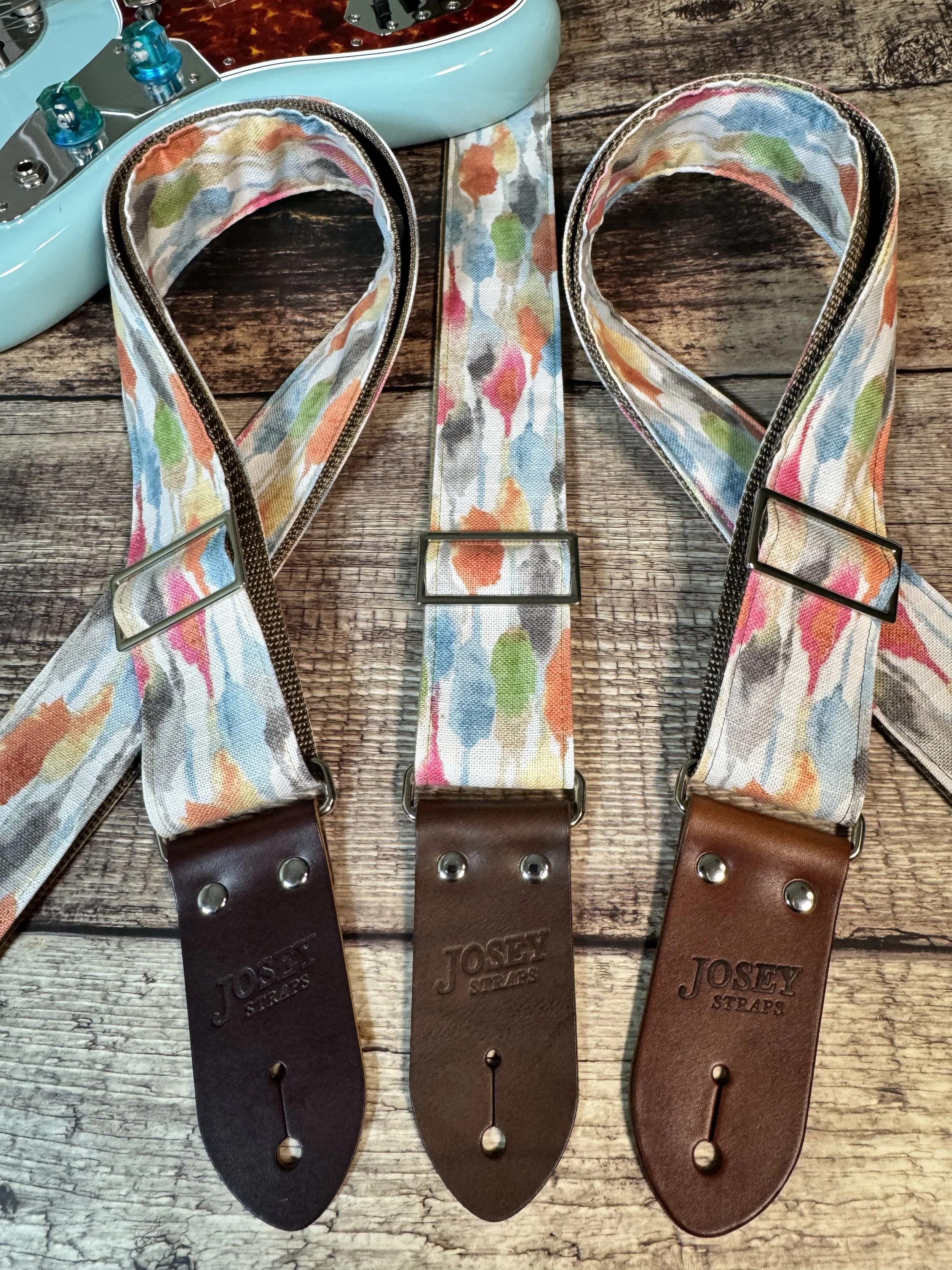 Josey Straps guitar straps in the April colorway — a watercolor print of coral, blue, sage, and amber on cream — displayed on a wood surface with a daphne blue Fender guitar.