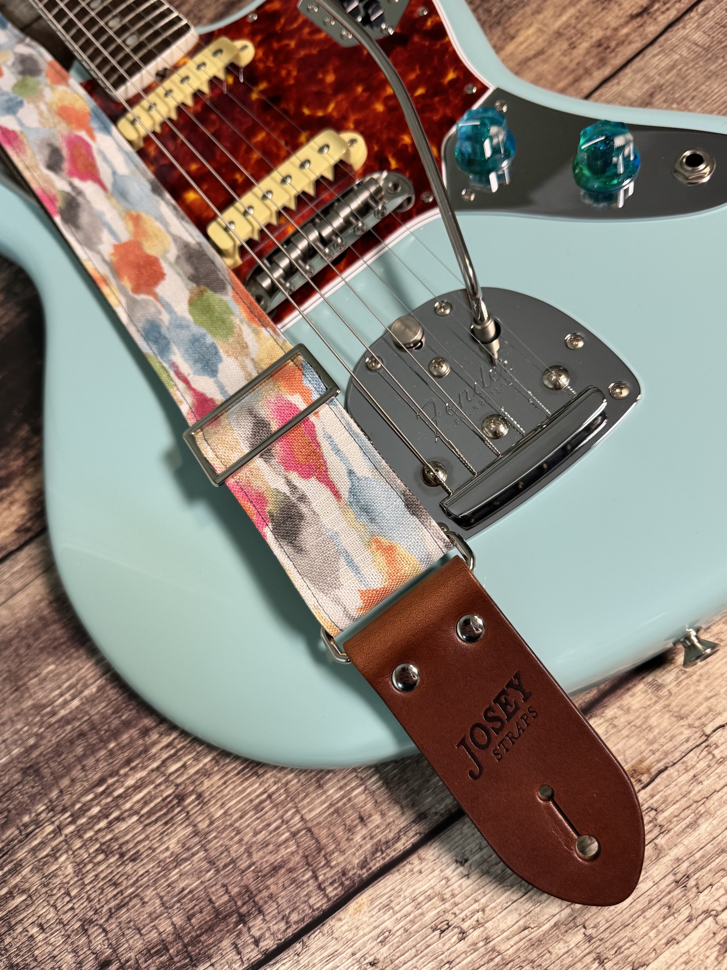 Josey Straps guitar straps in the April colorway — a watercolor print of coral, blue, sage, and amber on cream — displayed on a wood surface with a daphne blue Fender guitar.