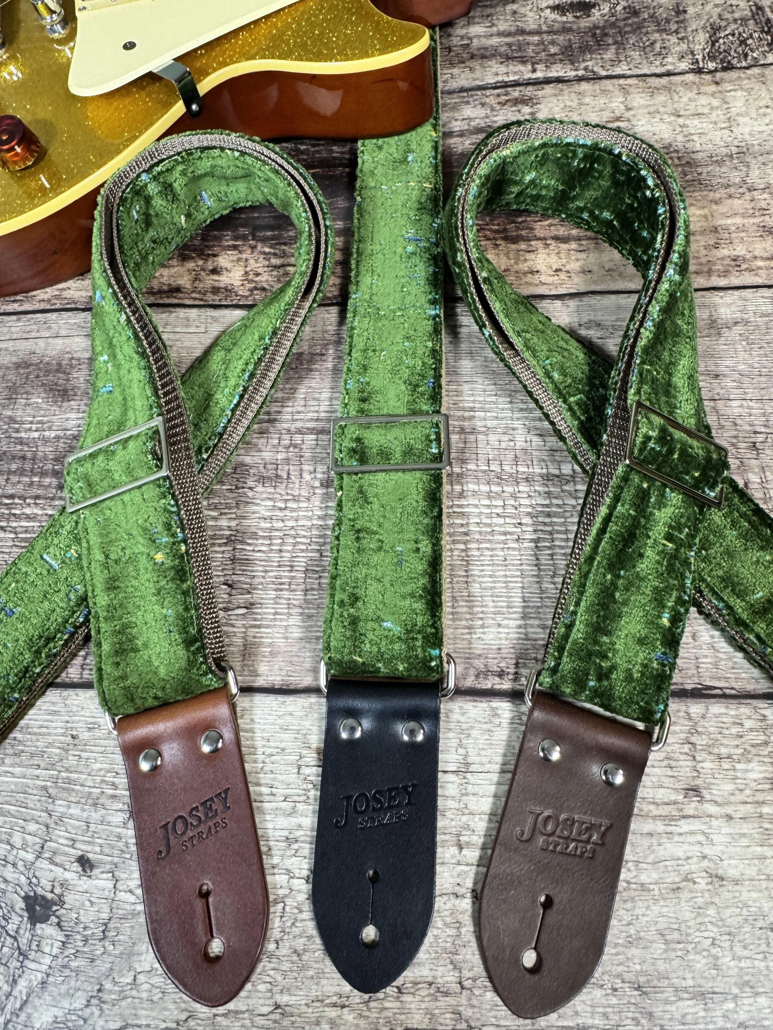 Handmade green velvety guitar strap with stamped leather ends laid on weathered wood next to a gold sparkle electric guitar — The Grouch by Josey Straps