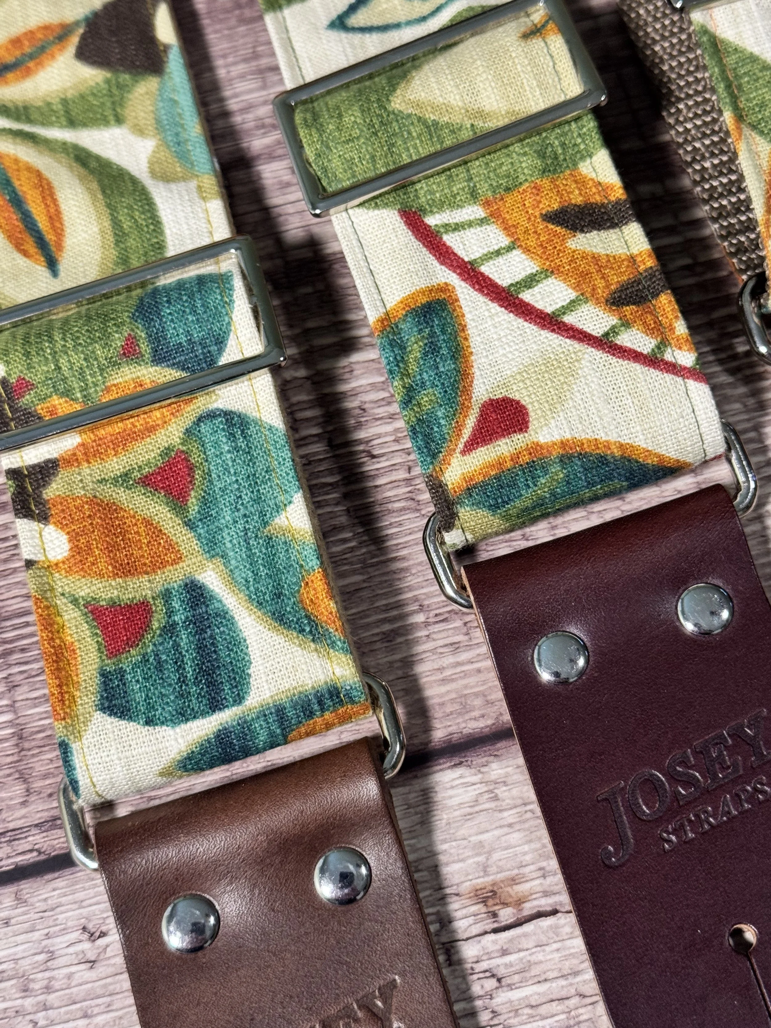 Handmade guitar strap in cream botanical fabric with amber, teal, burgundy, and forest green floral print, laid flat on a wood surface.