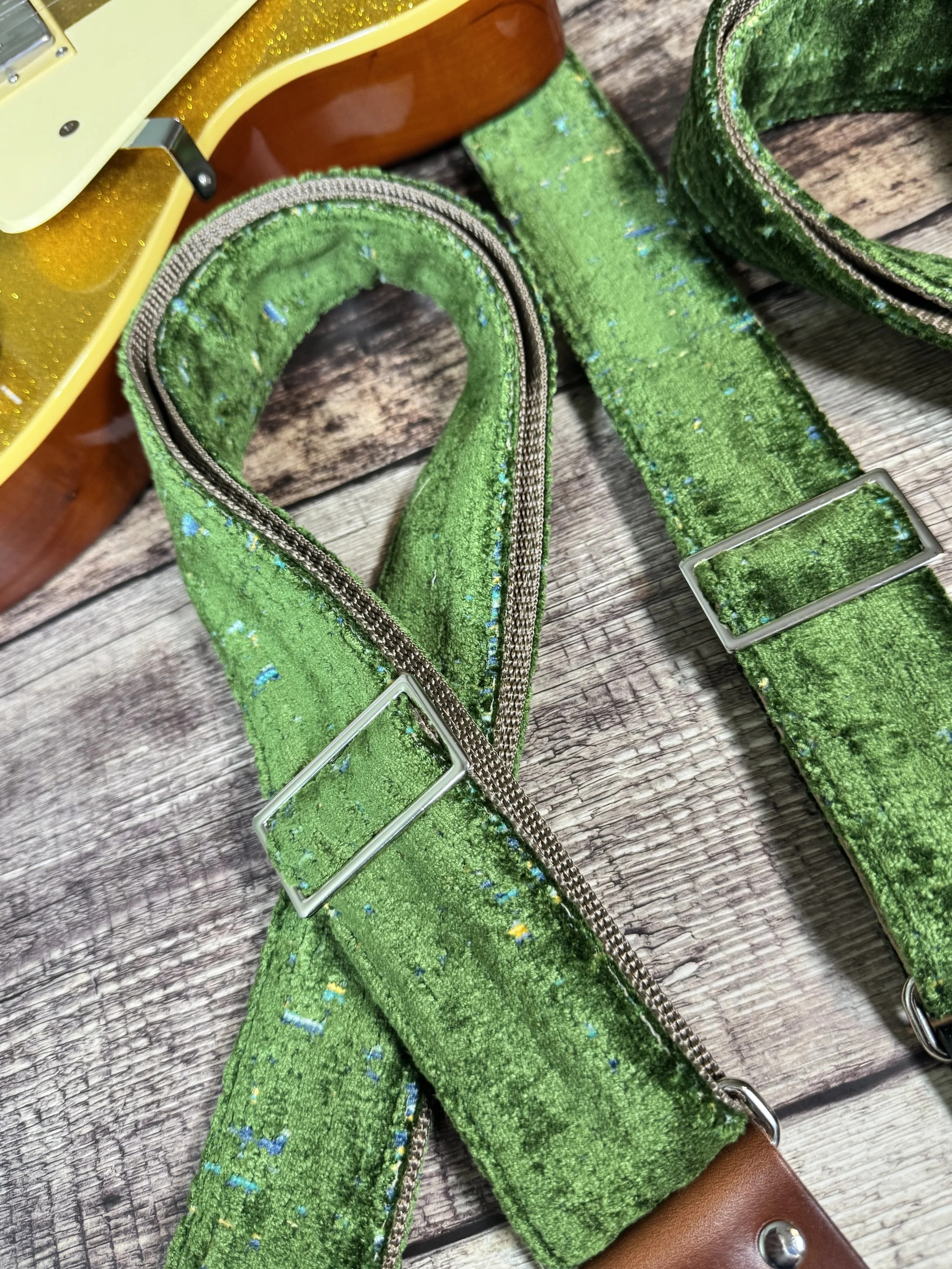 Handmade green velvety guitar strap with stamped leather ends laid on weathered wood next to a gold sparkle electric guitar — The Grouch by Josey Straps
