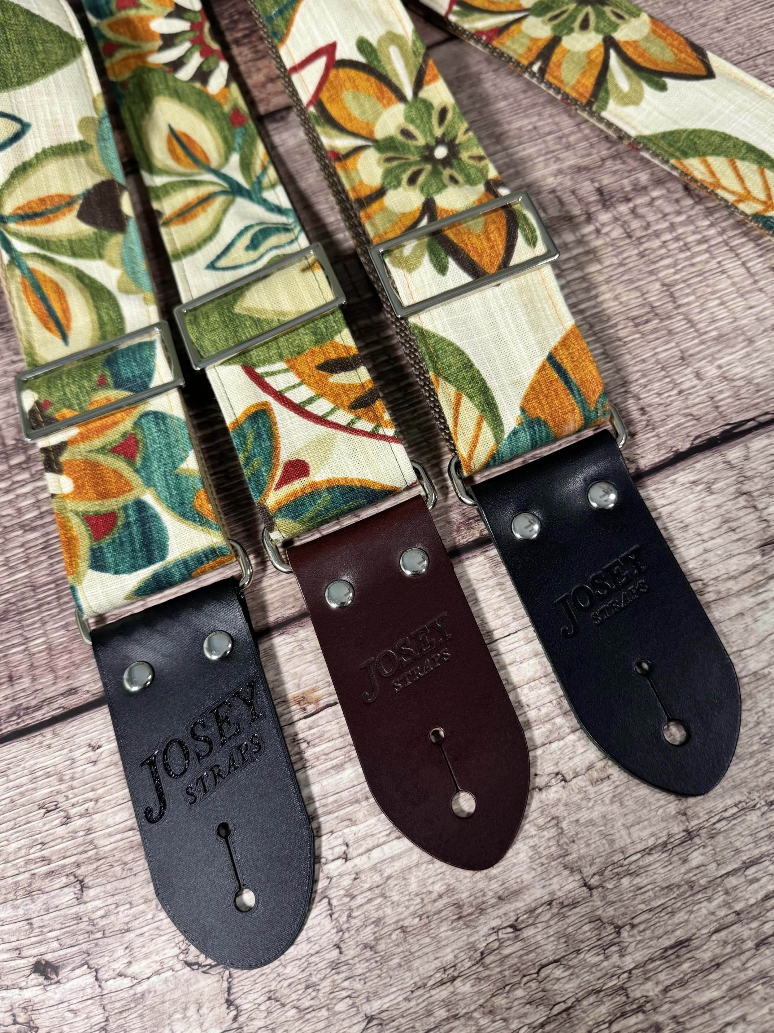 Handmade guitar strap in cream botanical fabric with amber, teal, burgundy, and forest green floral print, laid flat on a wood surface.