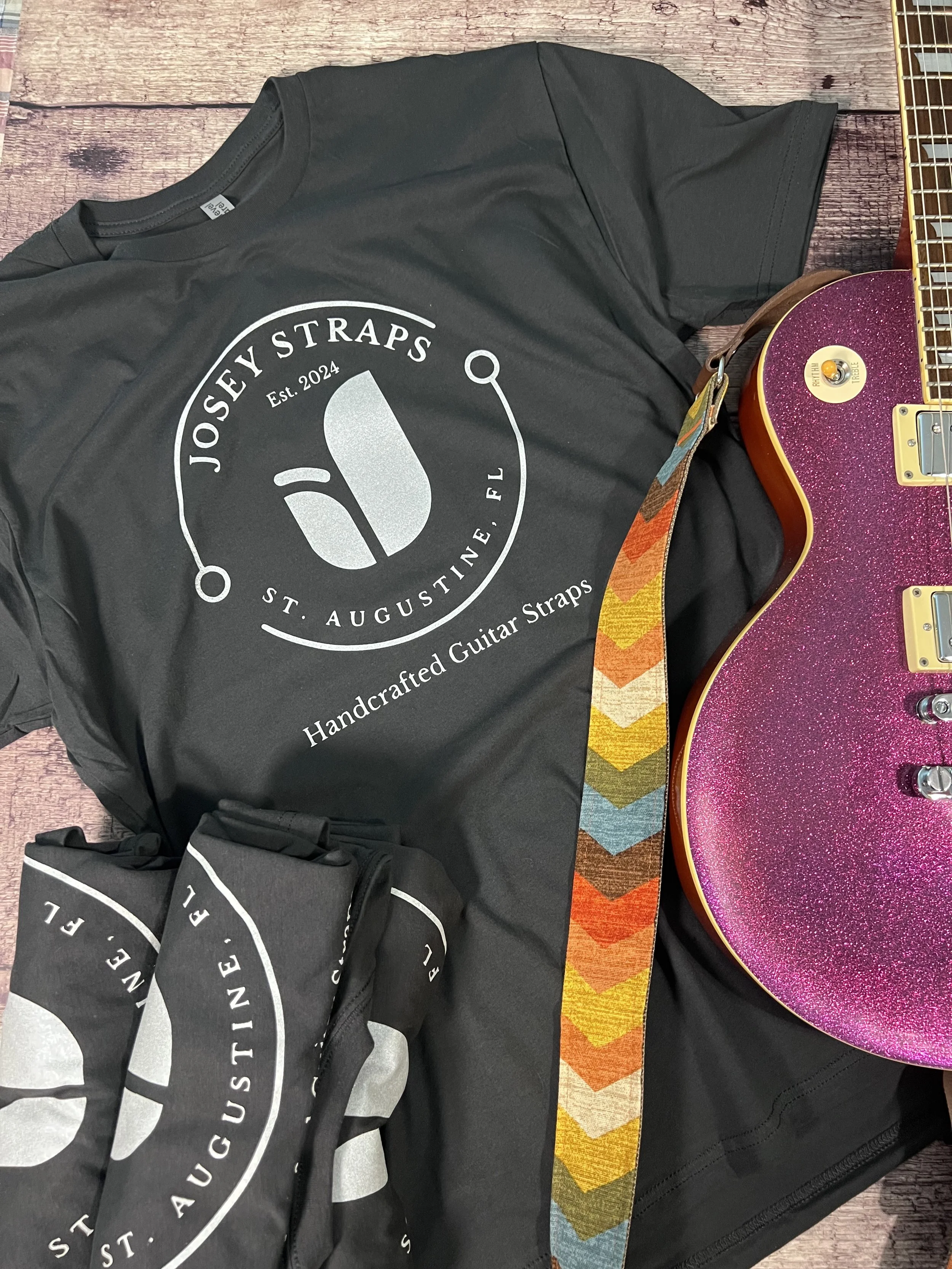 Vintage black Josey Straps screen printed t-shirts paired with a purple guitar and arranged on a wooden floor backdrop.