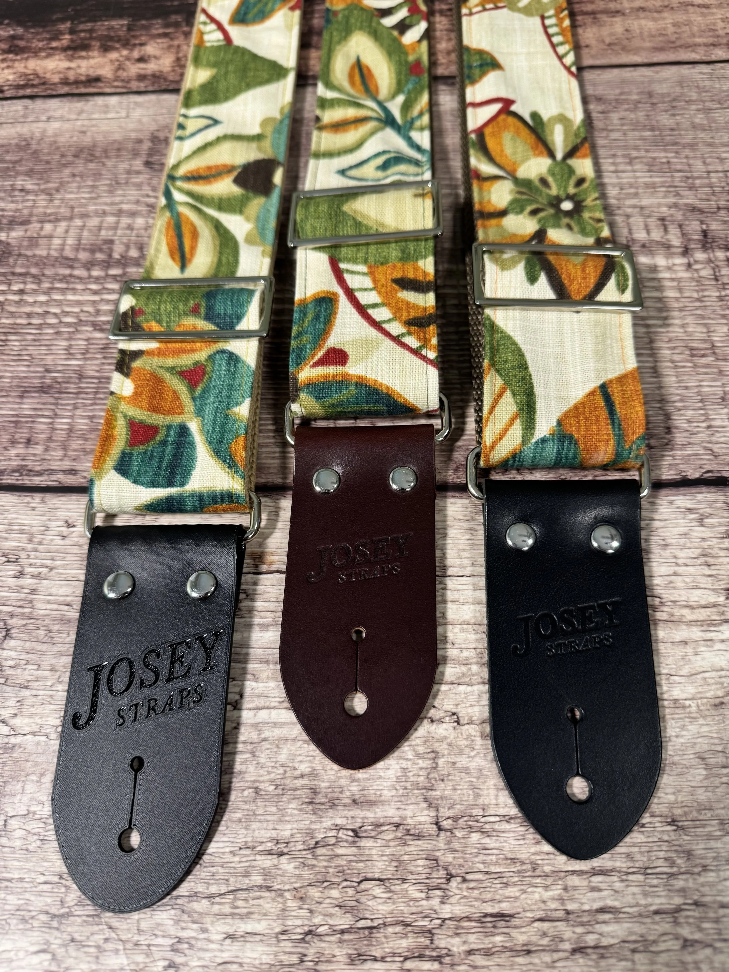 Handmade guitar strap in cream botanical fabric with amber, teal, burgundy, and forest green floral print, laid flat on a wood surface.