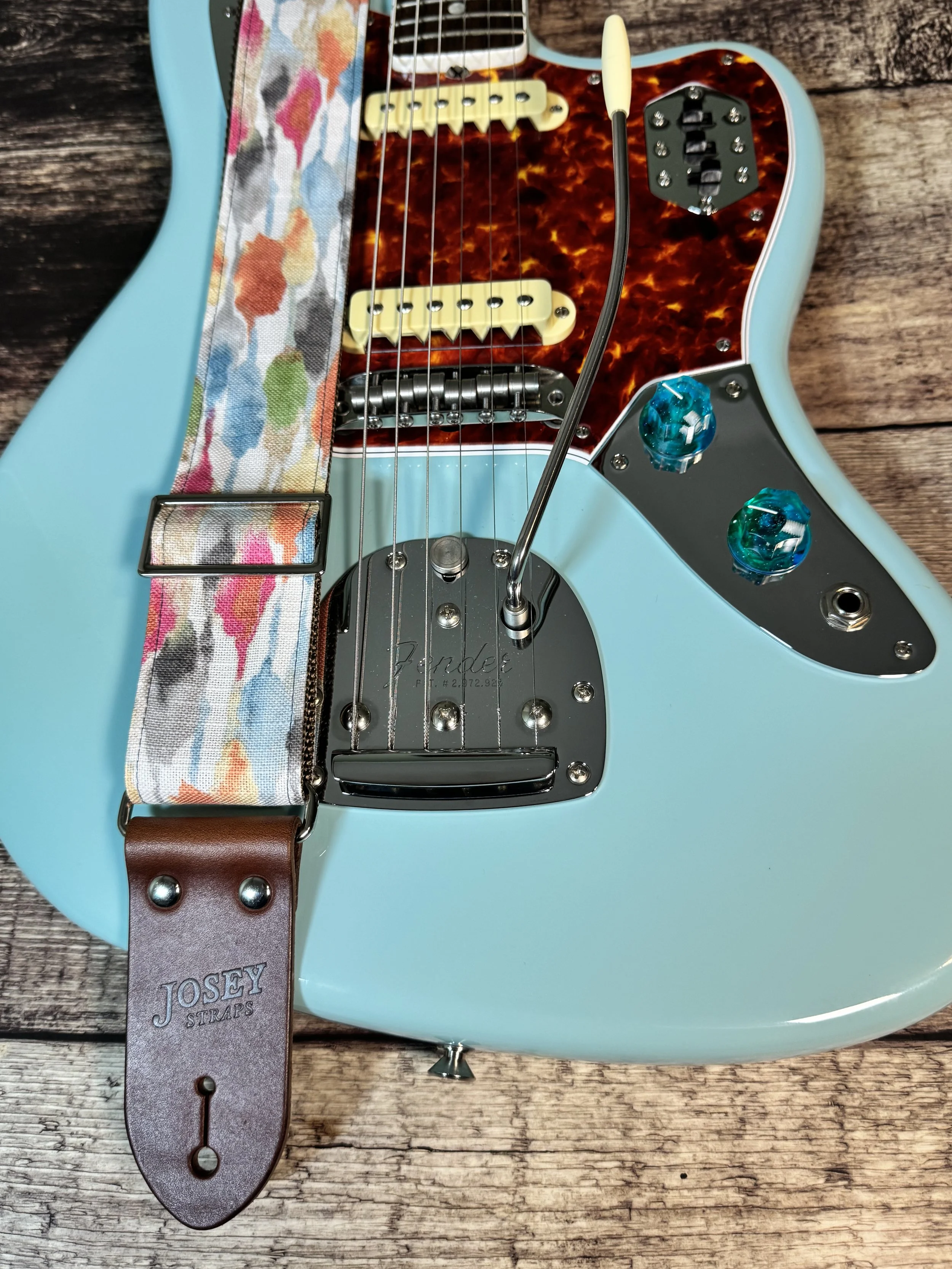 Josey Straps guitar straps in the April colorway — a watercolor print of coral, blue, sage, and amber on cream — displayed on a wood surface with a daphne blue Fender guitar.